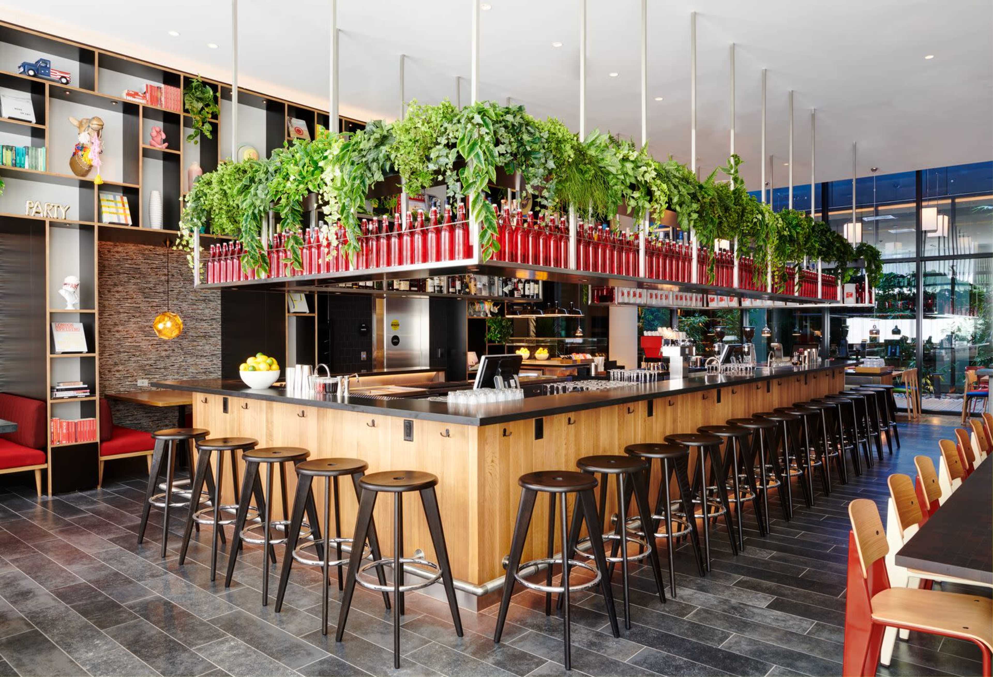 A modern bar area features a long wooden counter with high stools and a design accentuated by vibrant greenery overhead.