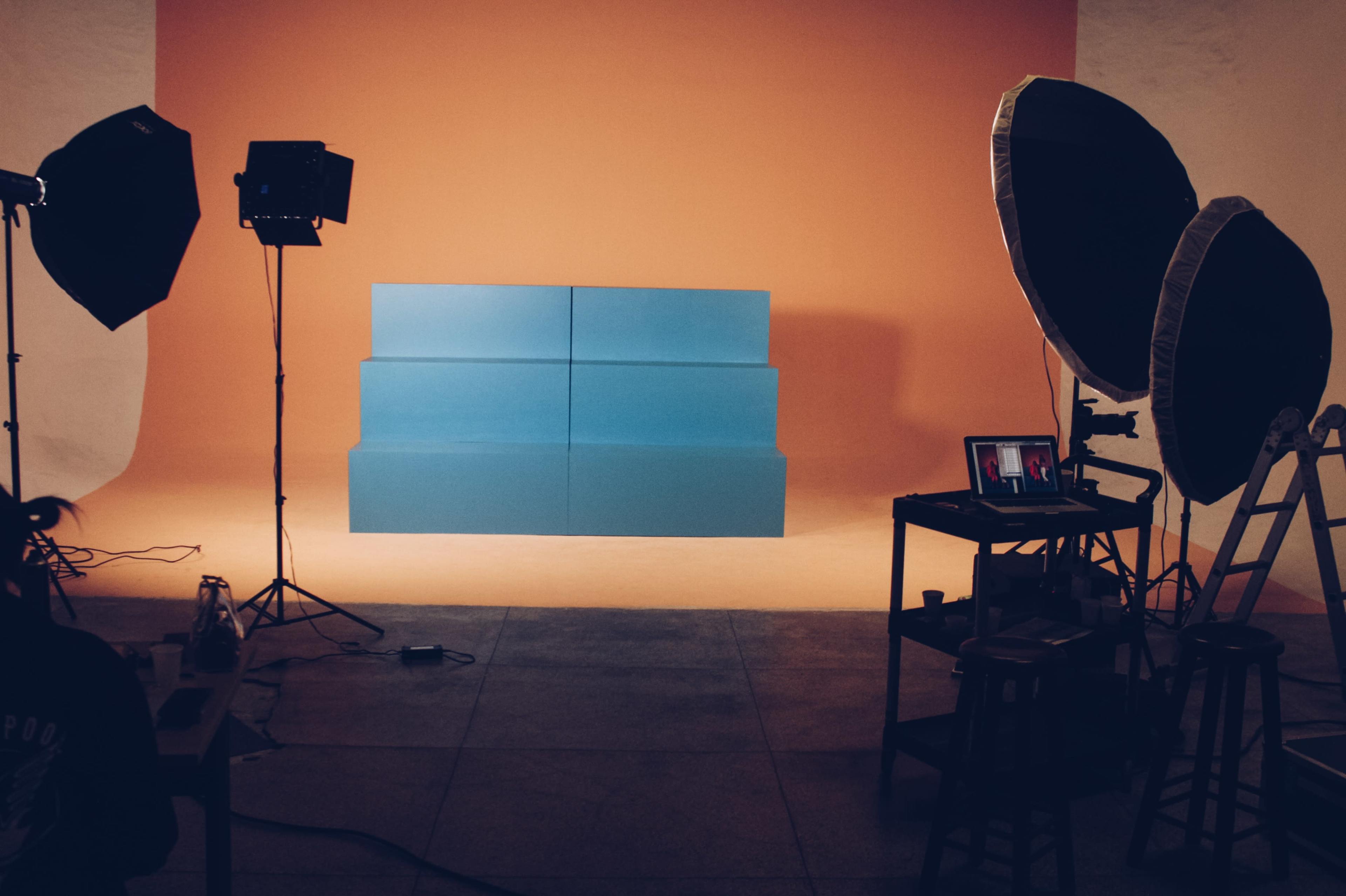 The image shows a photography studio setup with vibrant orange and blue backdrops, softbox lighting, and a table with equipment.