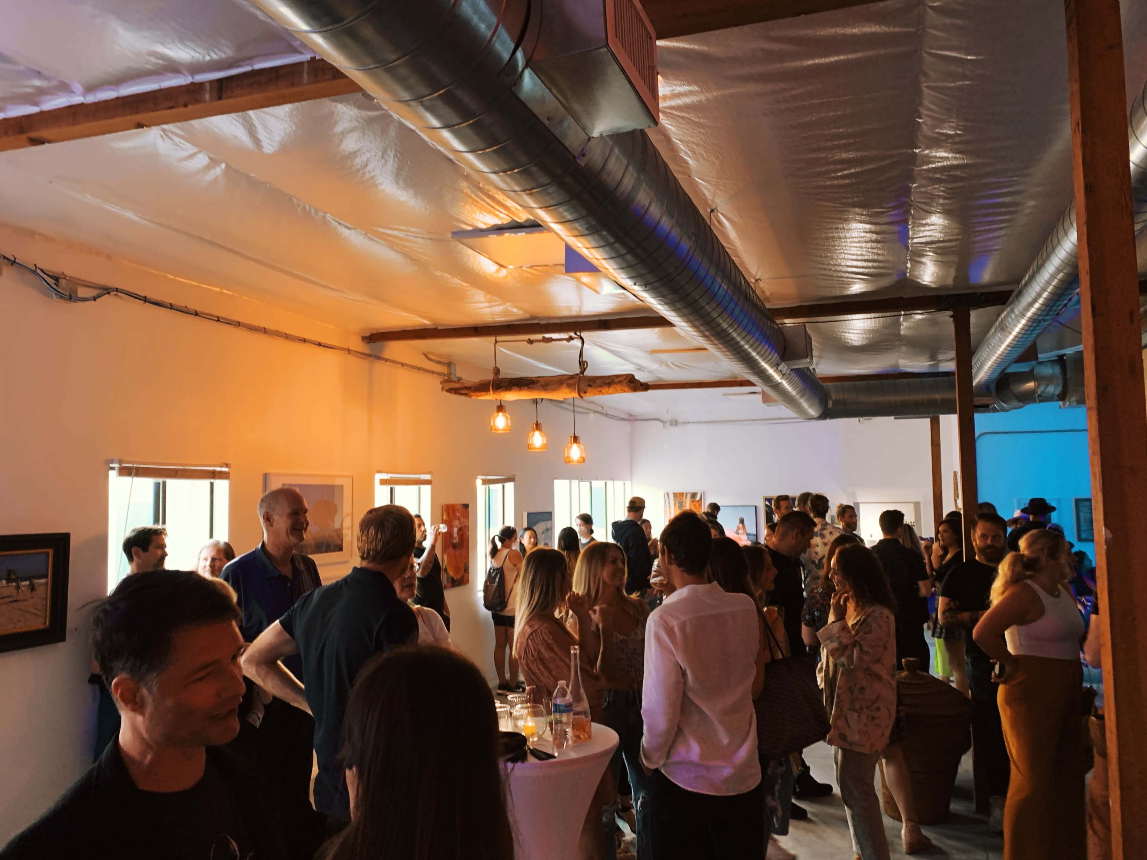 A crowded indoor space featuring people socializing and engaging at a gathering, with exposed beams and hanging lights.