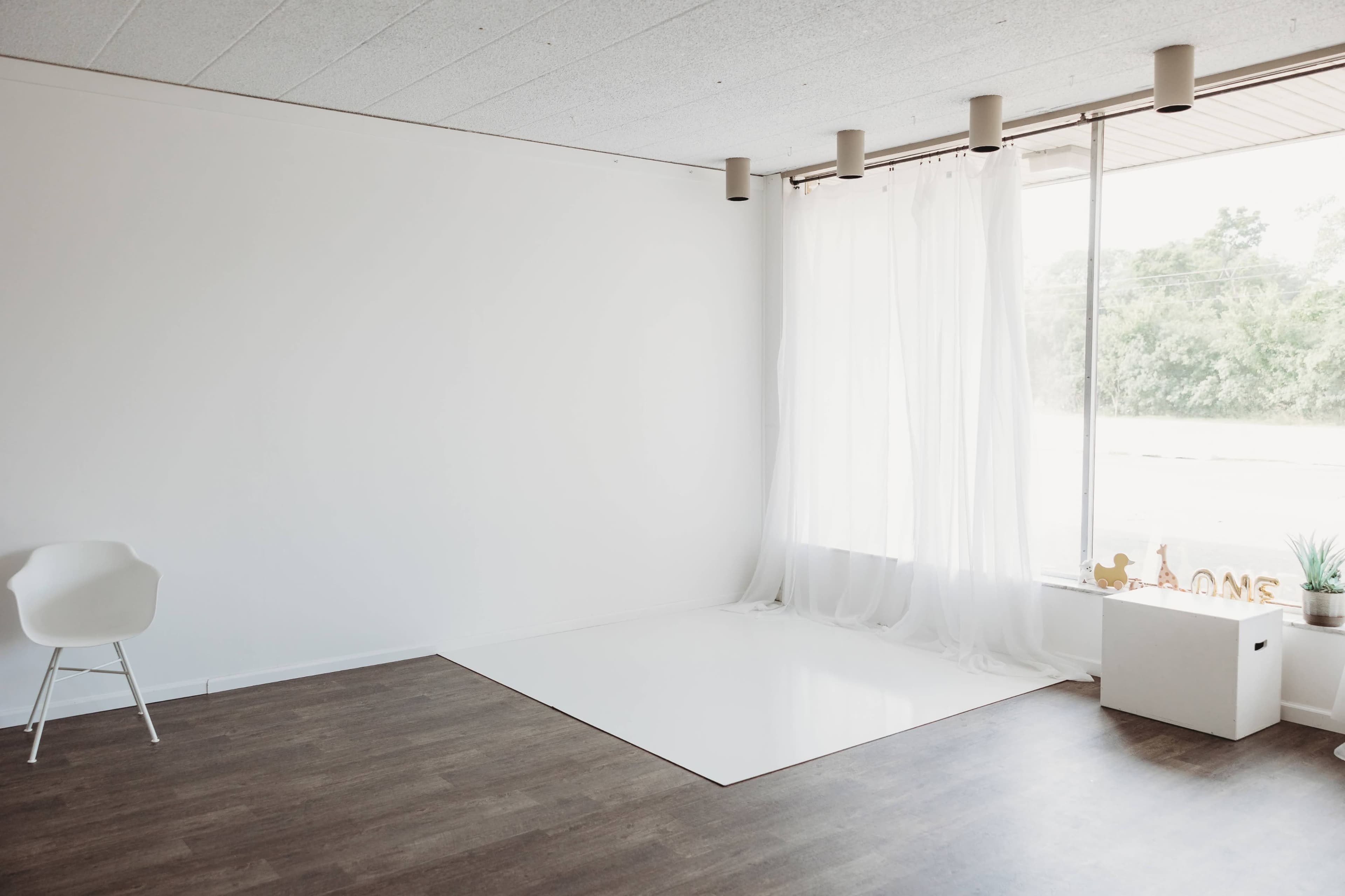 A minimalist room with white walls, a large window covered by sheer curtains, a white chair, and a small storage box on the floor.