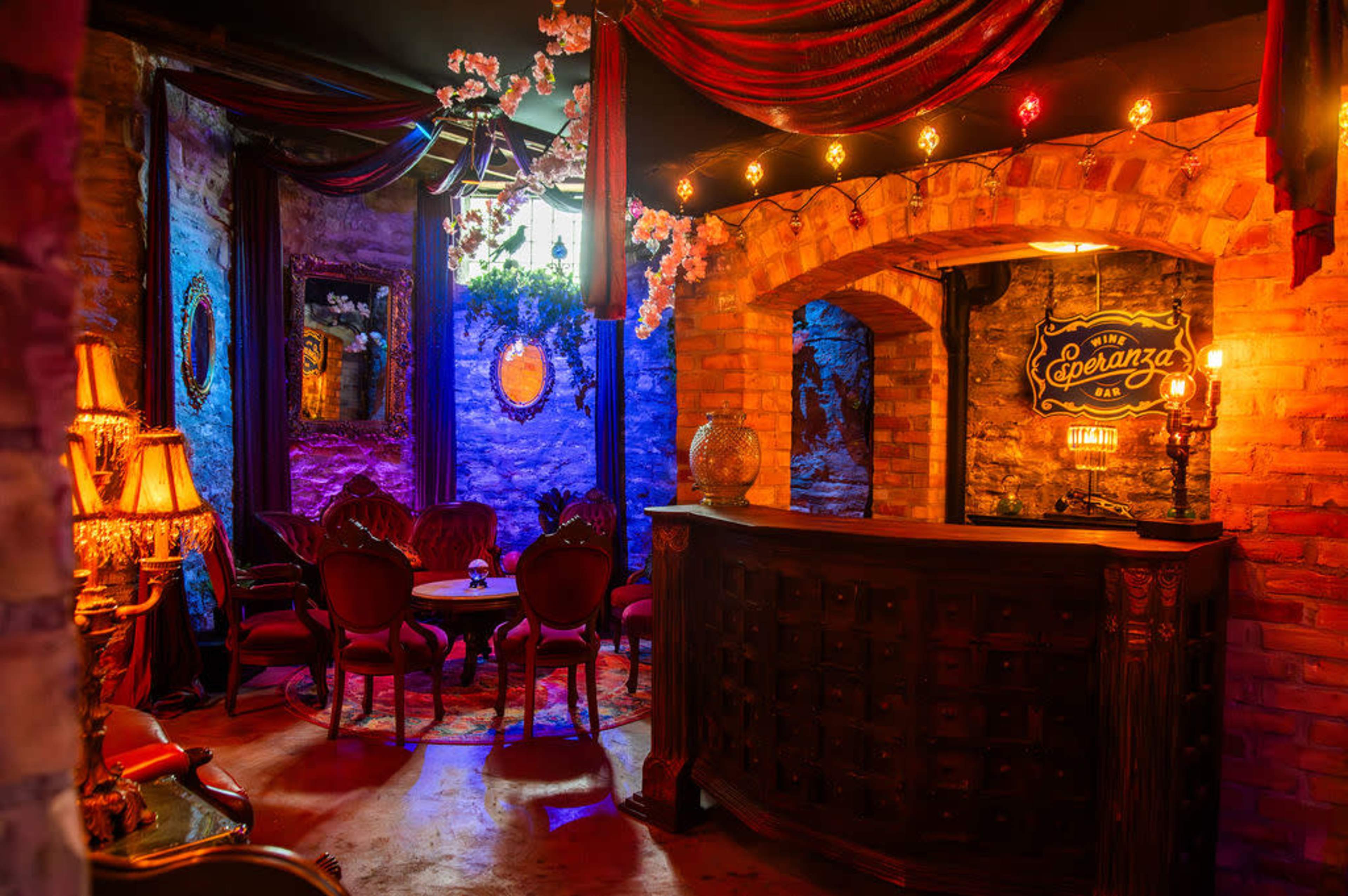 The image shows a cozy interior of a bar with a vintage-style wooden counter, red velvet seating, and colorful lighting illuminating the brick walls.