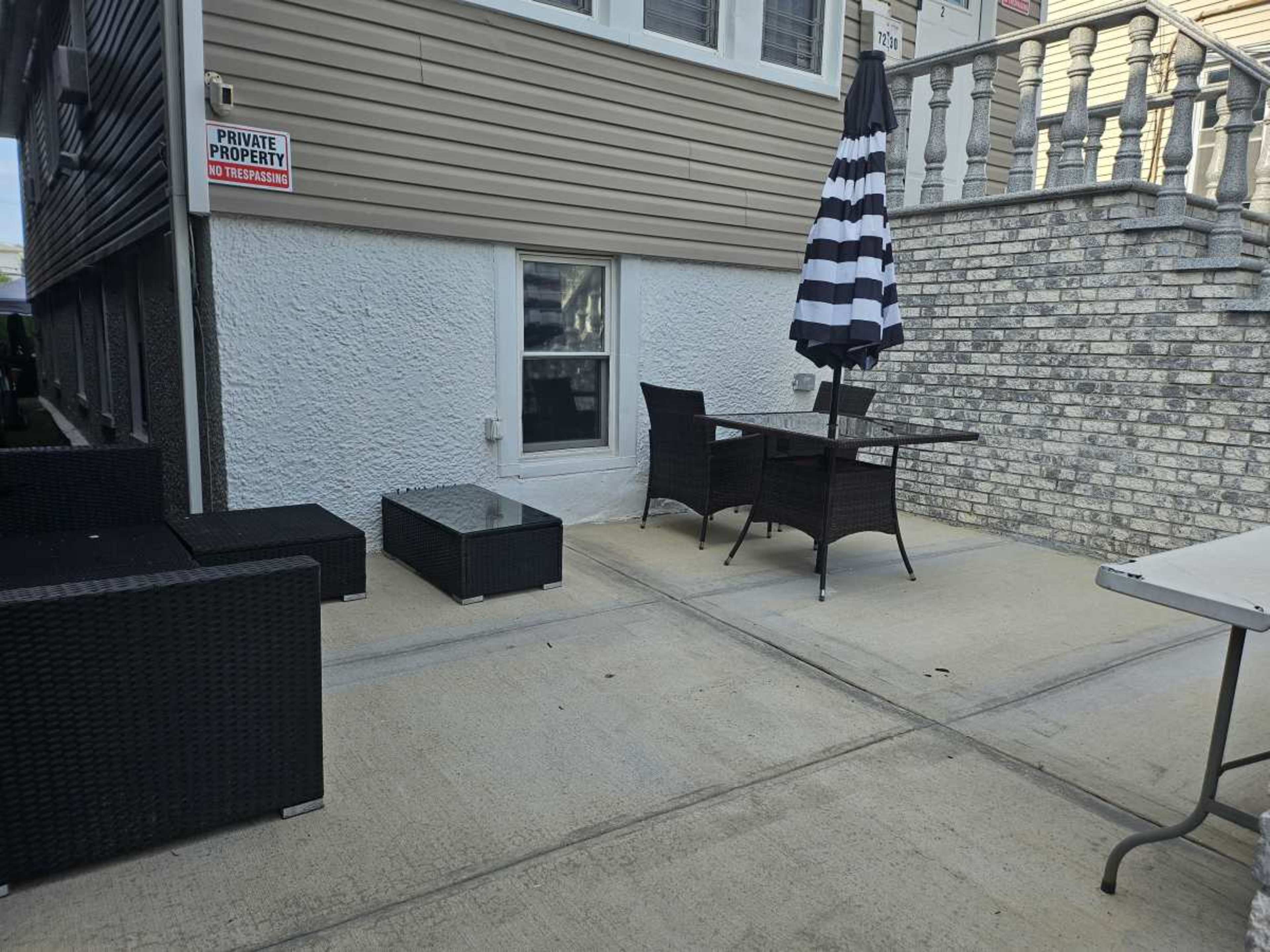 A small outdoor seating area with wicker furniture, a striped umbrella, and a concrete floor near a building.
