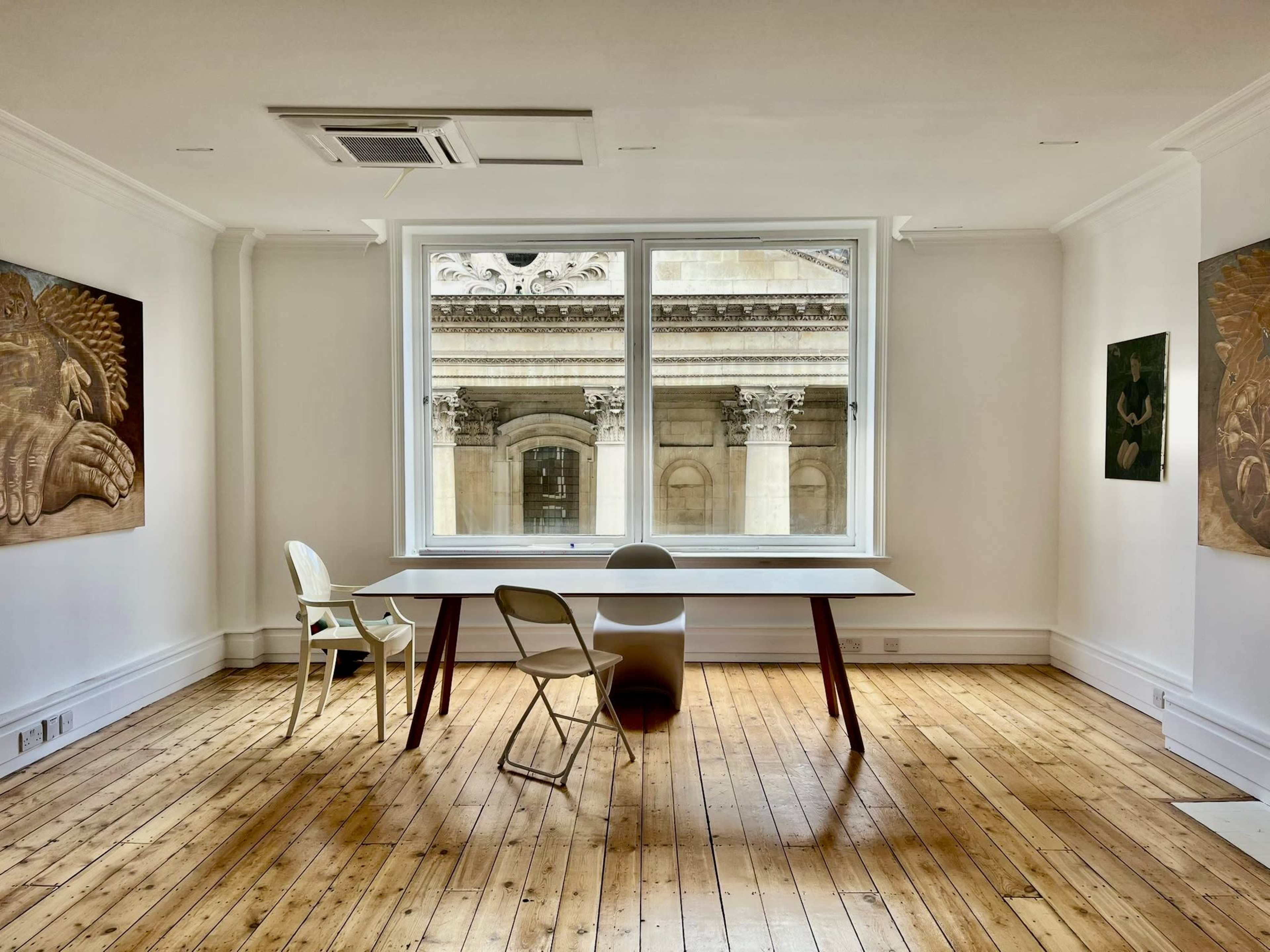 A minimalist room features a large window overlooking a historic facade, with a table and chairs arranged on wooden flooring.