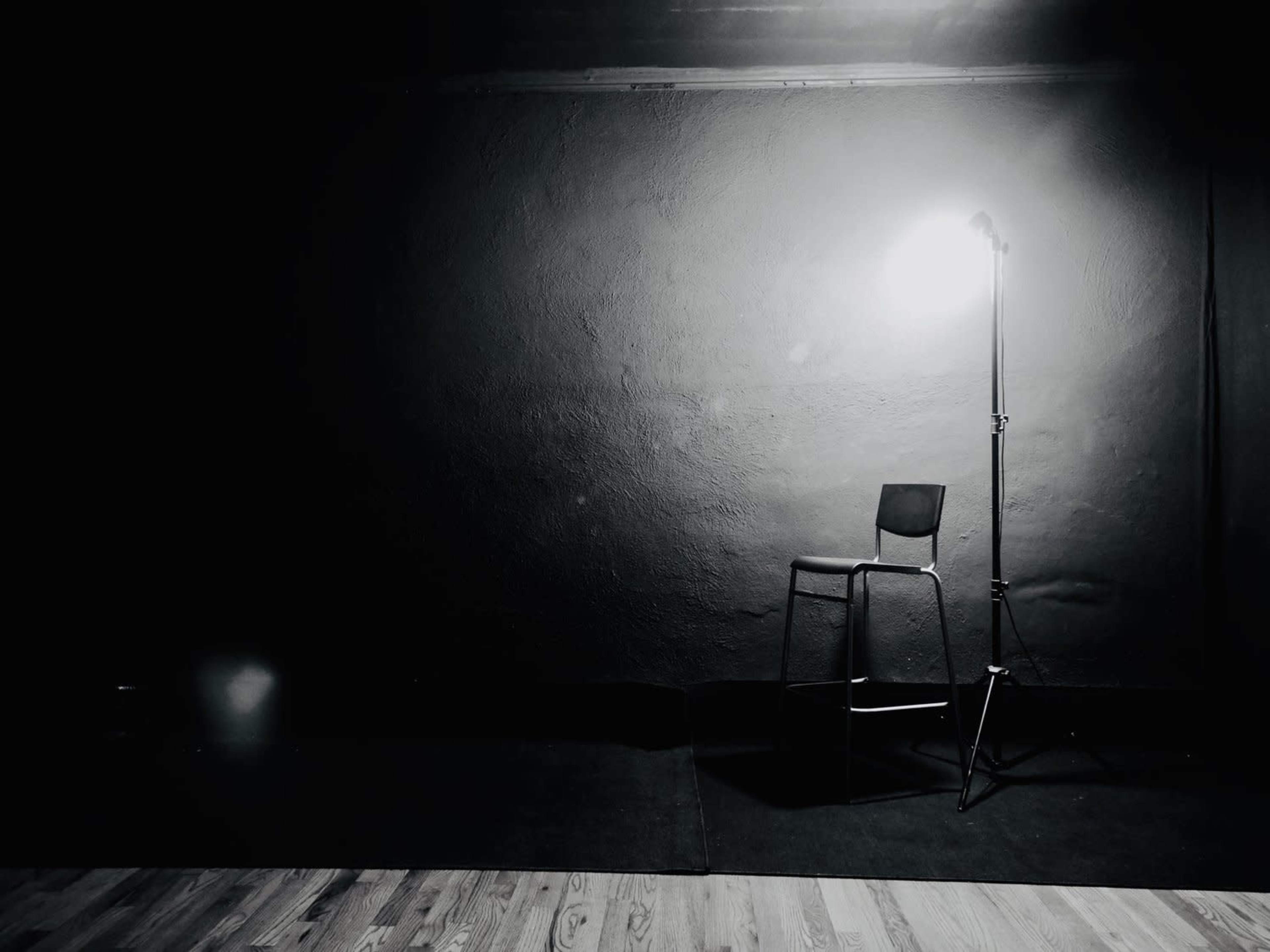 A single chair is positioned under a spotlight in a dimly lit room with a textured wall.