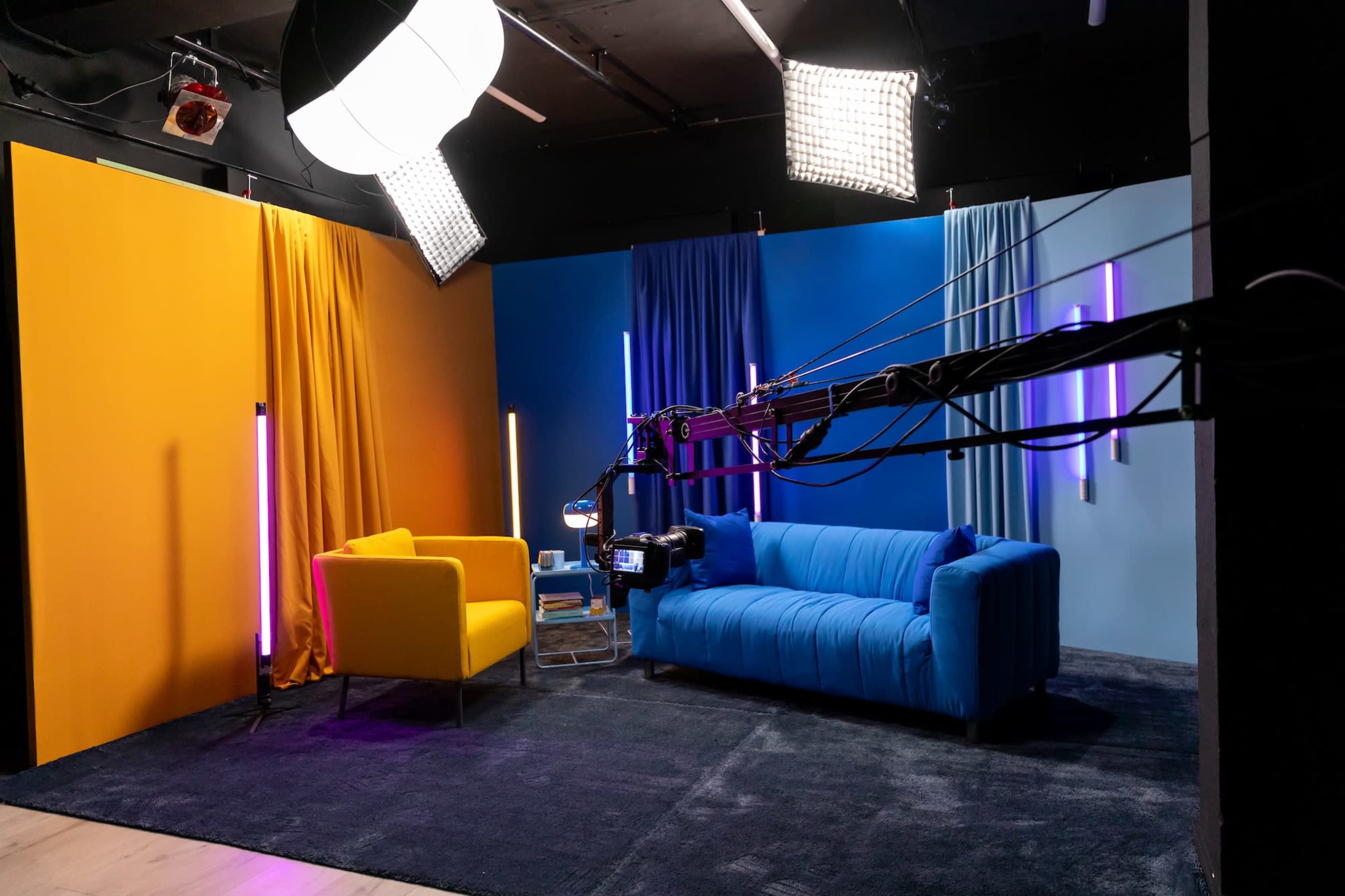 The image shows a brightly colored television studio setup with a yellow chair, a blue sofa, and vertical light strips against vibrant blue and orange backdrops.