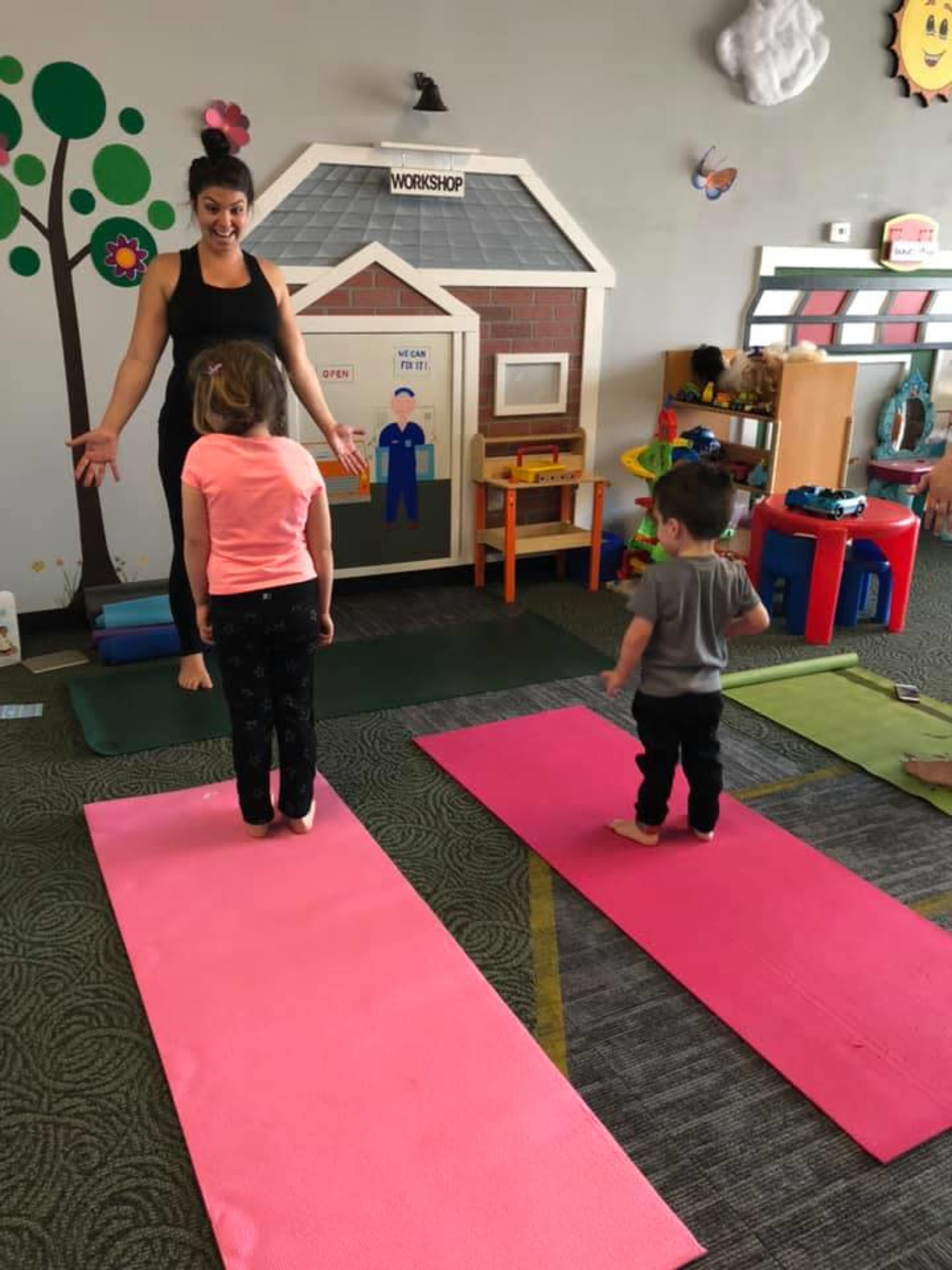 A woman demonstrates a yoga pose to two young children on colorful mats inside a playroom.
