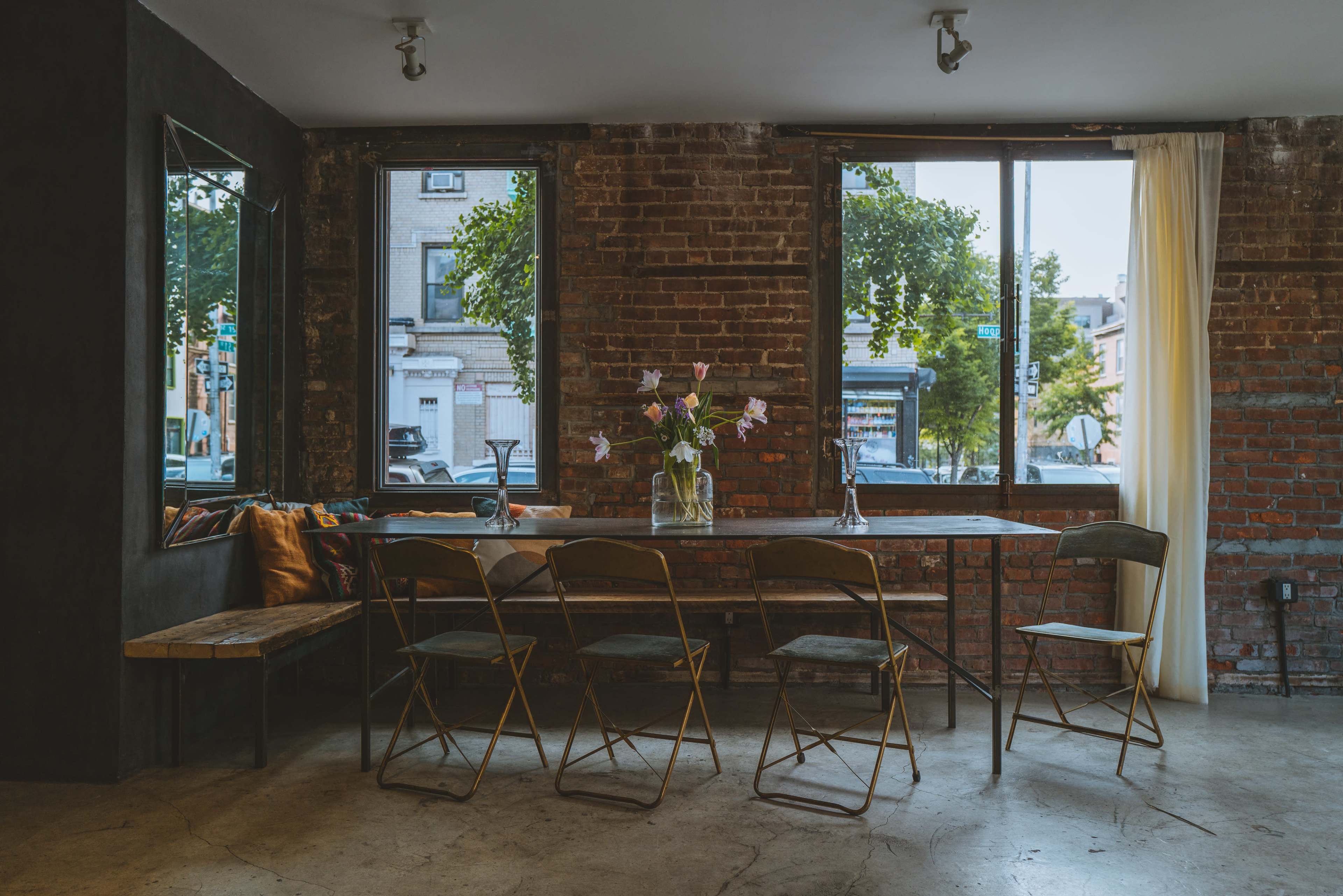 A long table with gold chairs sits in a room featuring exposed brick walls and large windows that let in natural light, complemented by a vase of flowers at the center.