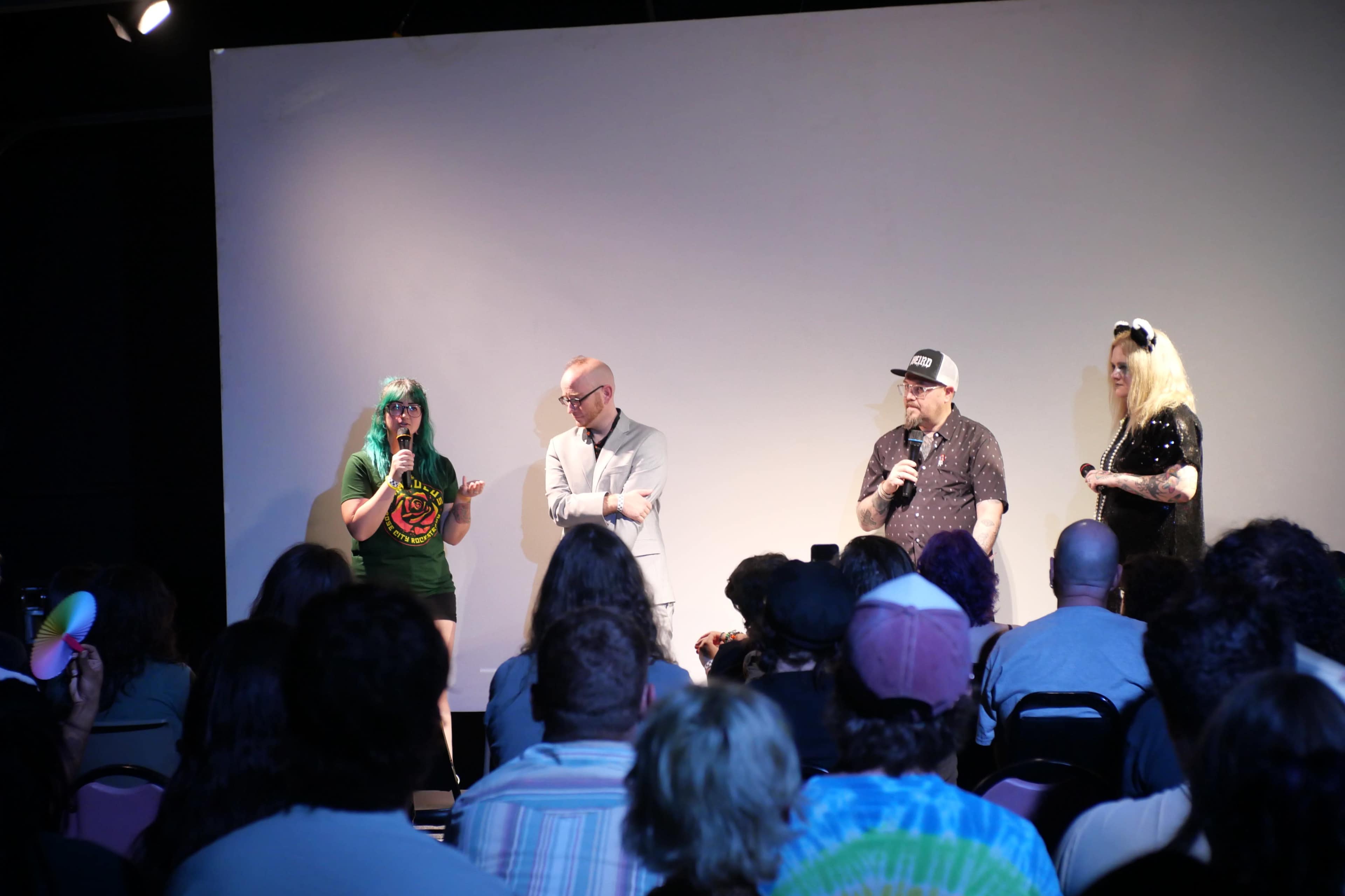 A panel discussion is taking place on stage with four speakers in front of an audience seated in a dimly lit room.