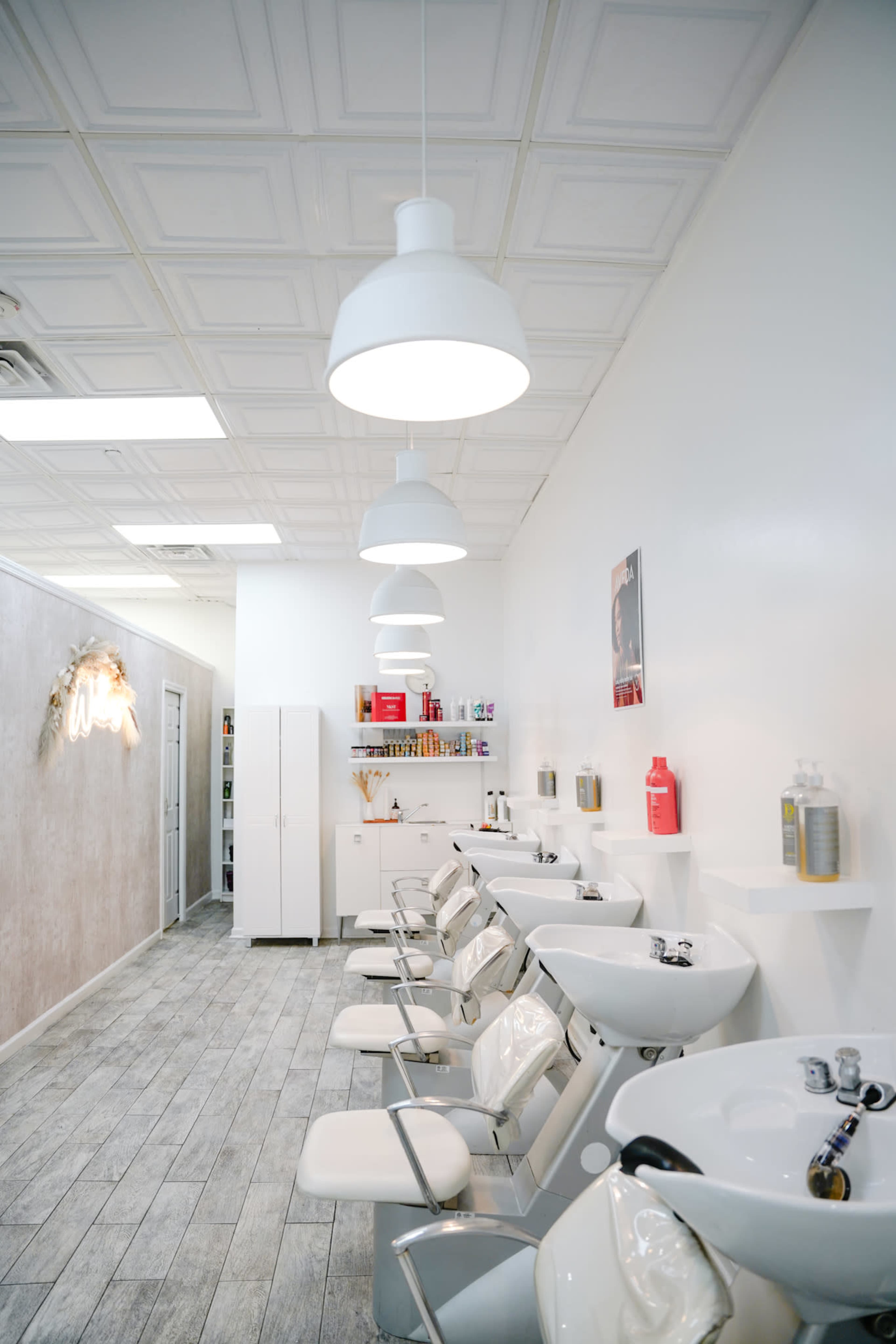 A bright hair salon features multiple styling stations with white sinks and modern lighting fixtures, along with storage cabinets and product displays along the walls.
