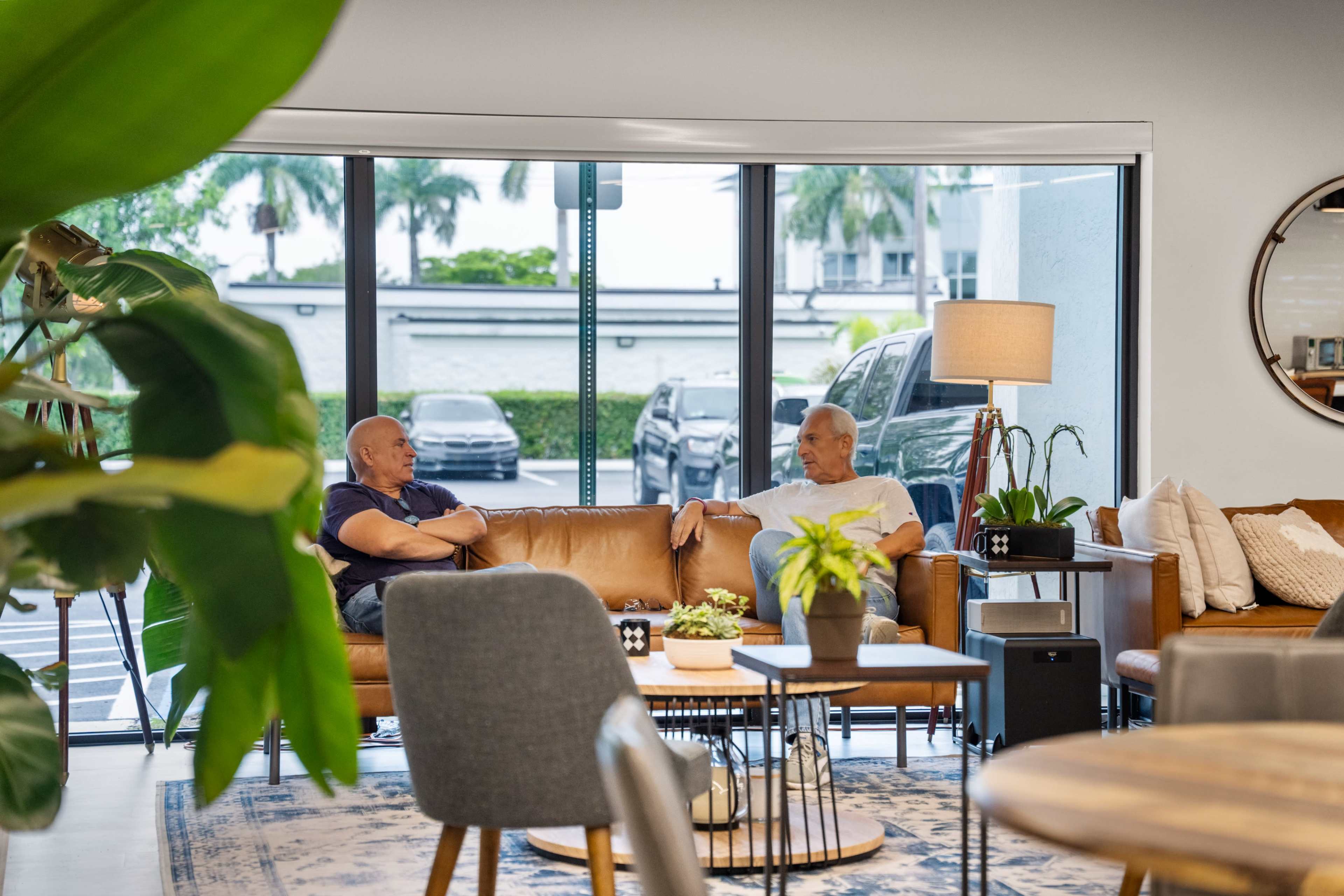 Two men are seated on a brown leather sofa in a modern lounge area, engaged in conversation, while large windows provide a view of parked cars and greenery outside.