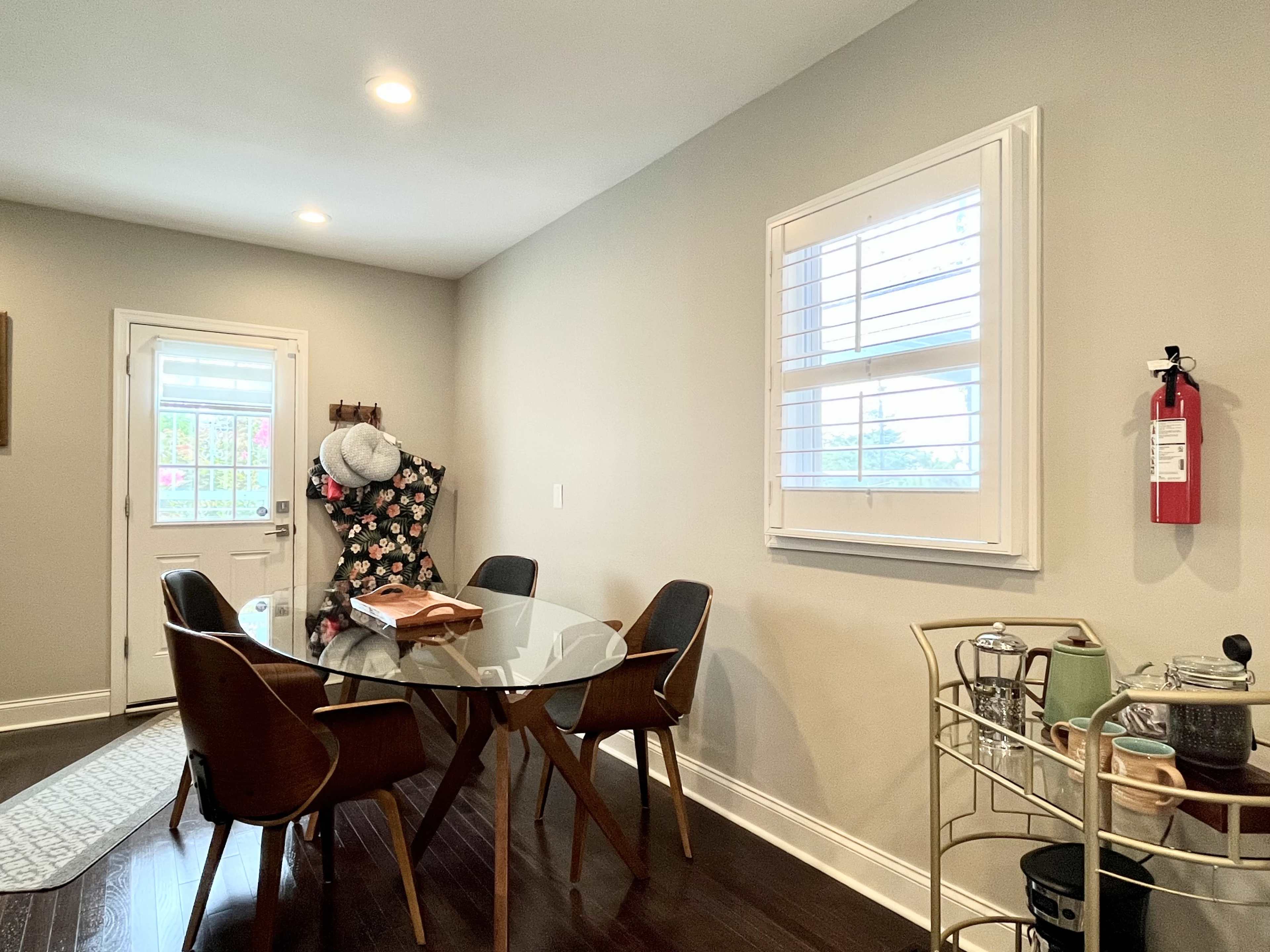 A dining area features a glass table surrounded by wooden chairs, with a door leading outside and a cart holding kitchen items.