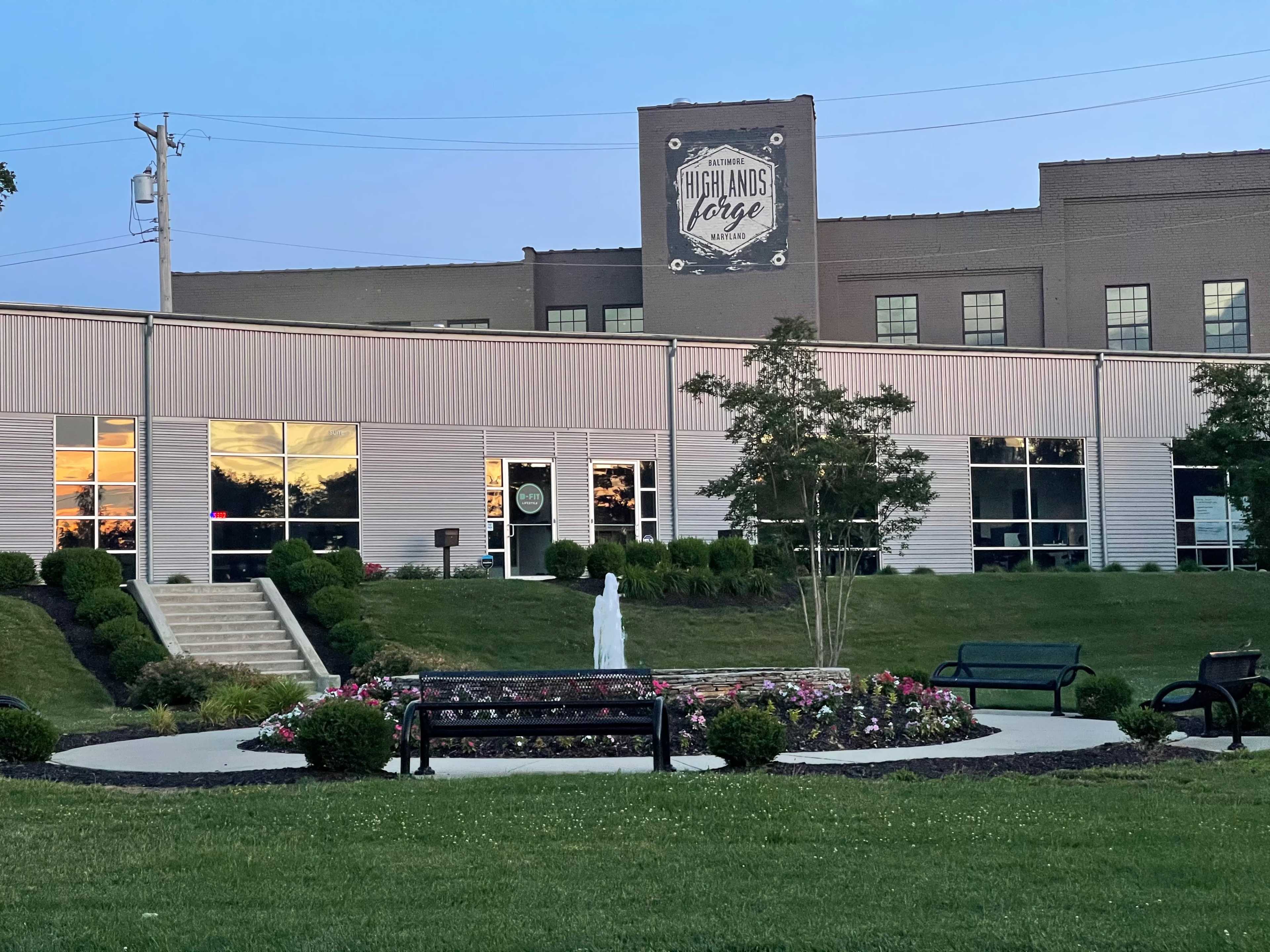 A modern building with large windows and a sign that reads "Highlands Forge," surrounded by landscaped gardens, a fountain, and benches in a grassy area during twilight.