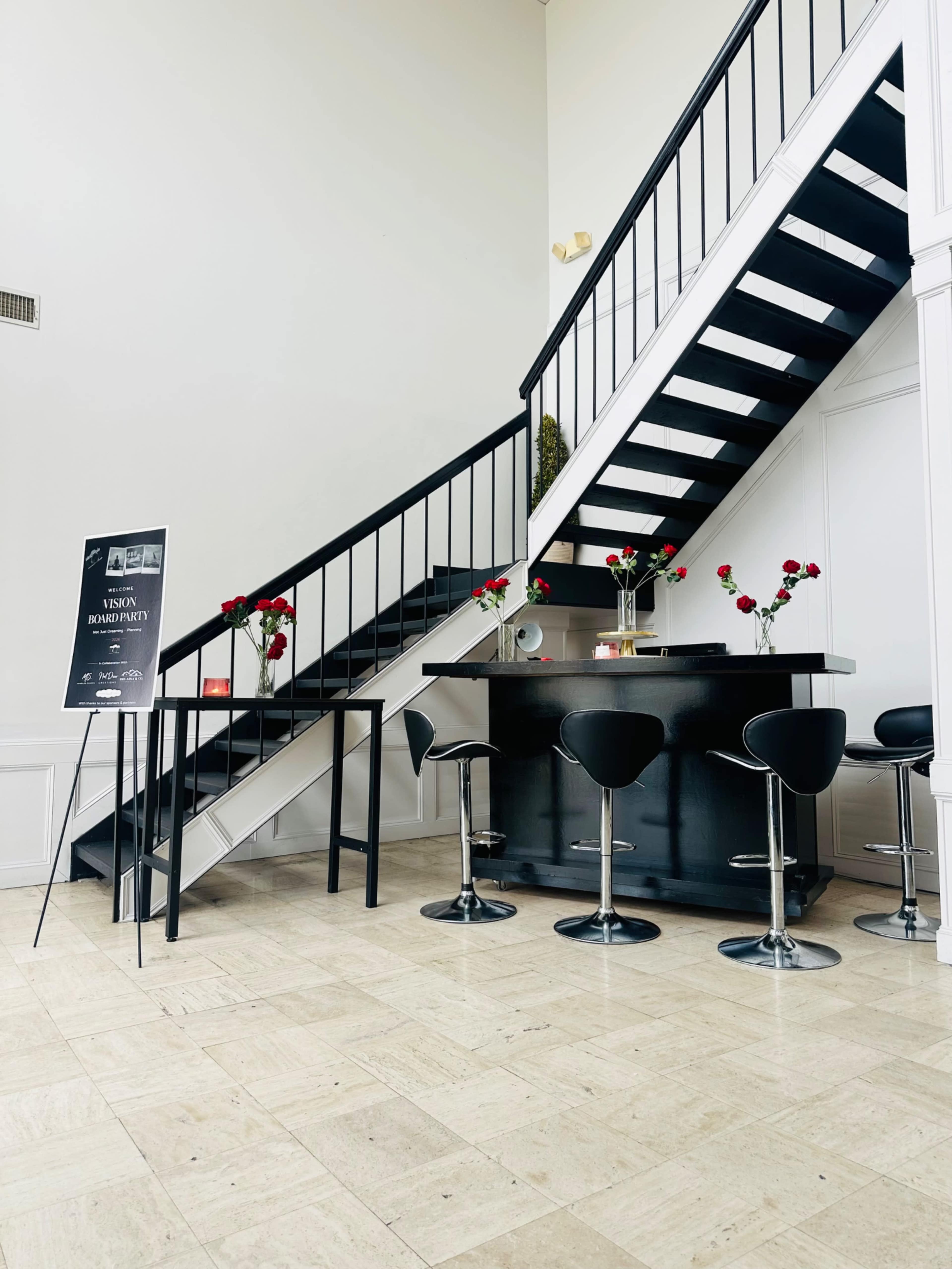 A modern interior features a bar with high stools beside a staircase, adorned with floral arrangements and a promotional sign.