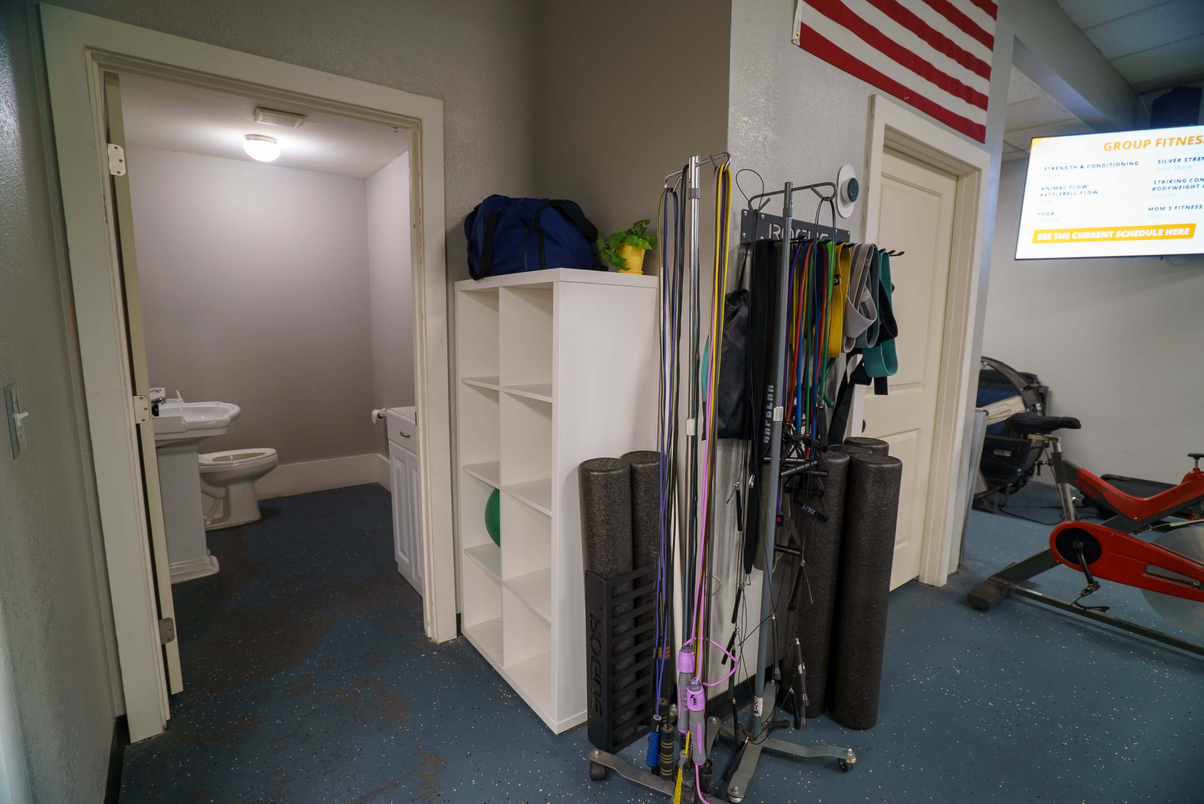The image shows a hallway leading to a bathroom with a toilet and a storage shelf, alongside fitness equipment and an American flag on the wall.