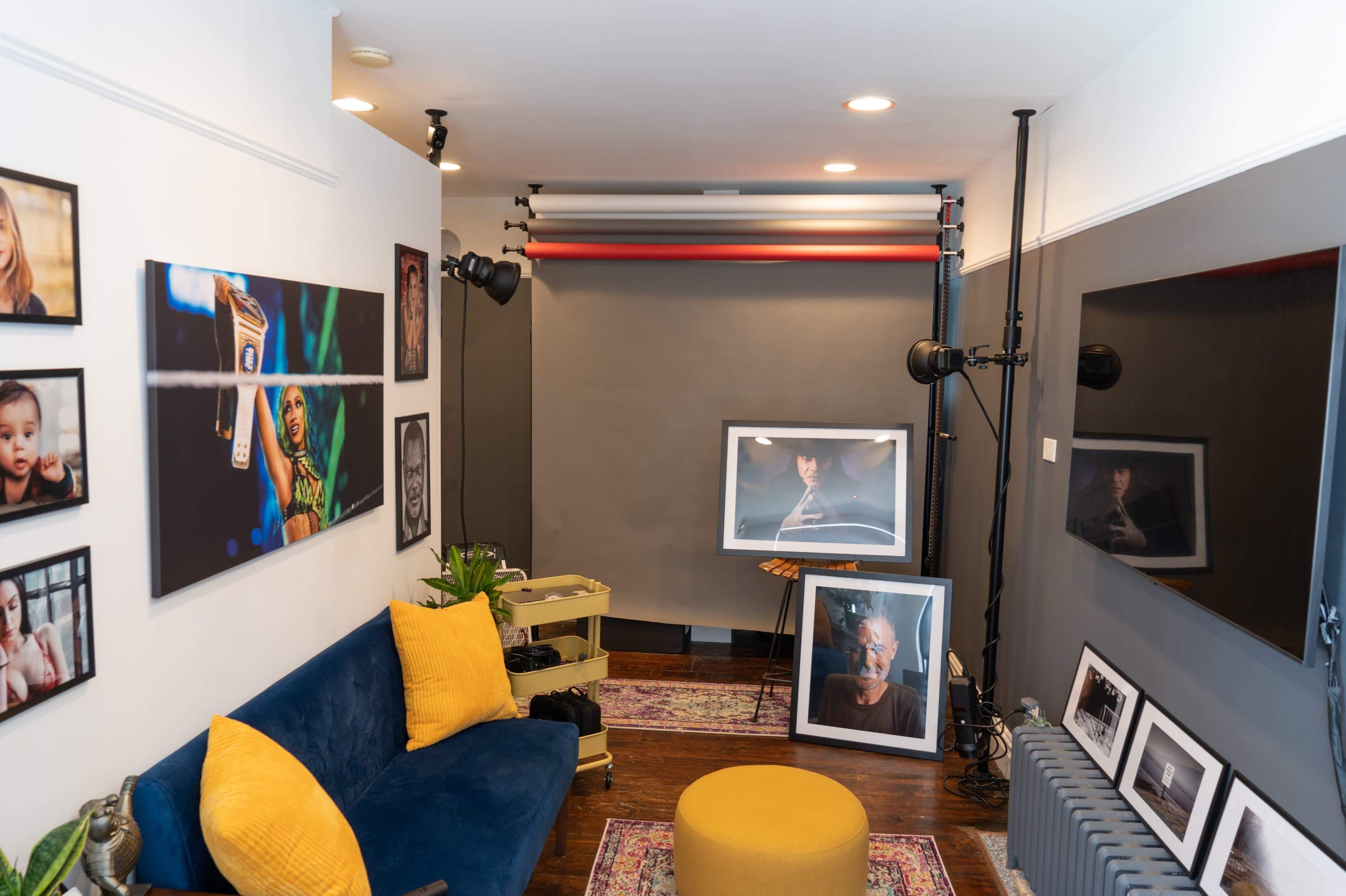 A small studio space featuring a blue sofa with yellow cushions, several framed photographs on the walls, a round ottoman, and a large flat-screen TV.
