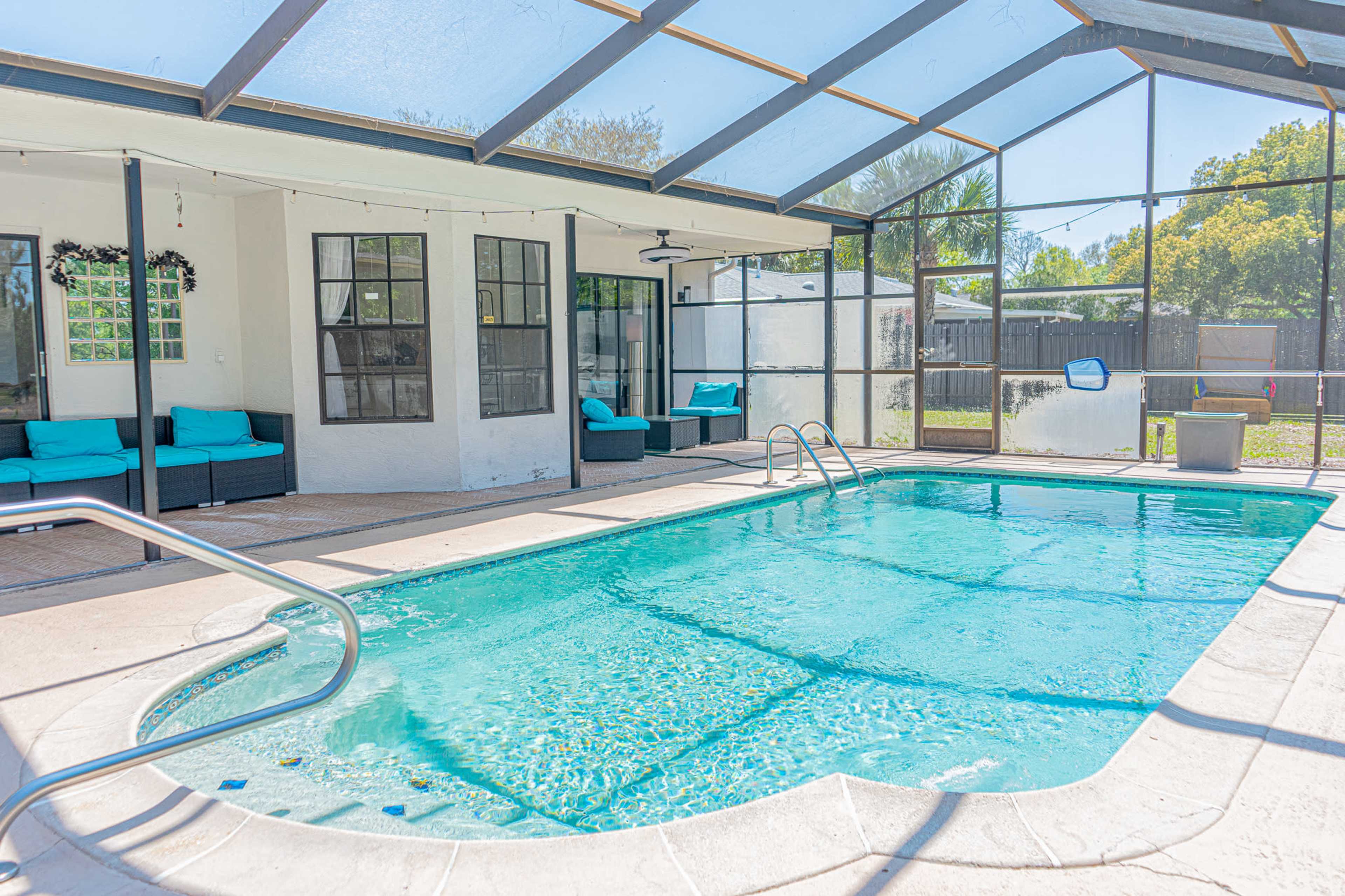 The image shows a screened-in pool area with a clear blue pool, surrounded by lounging furniture and a sunny outdoor view.
