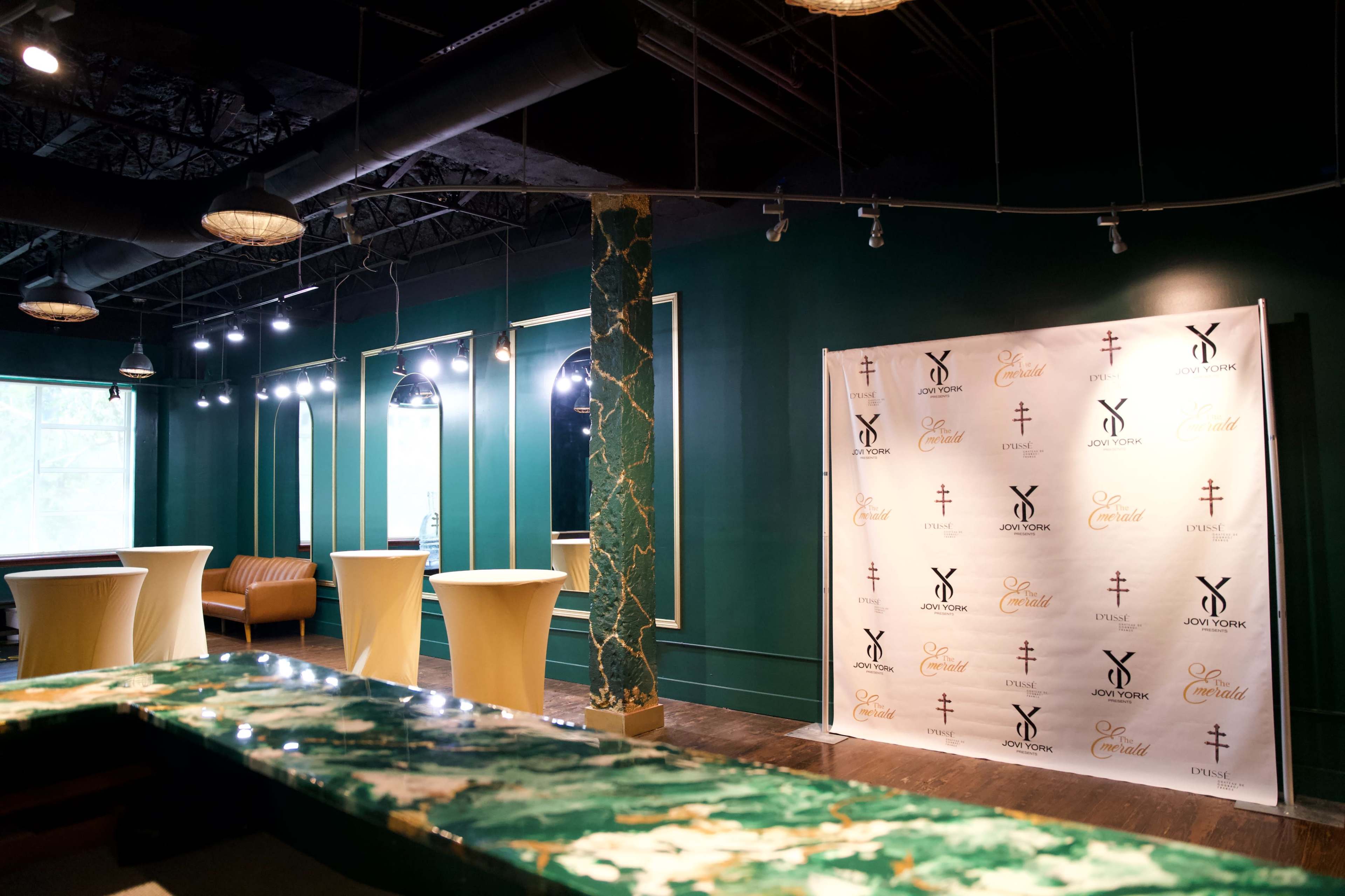 The image shows a vibrant interior space with green walls, elegant pendant lighting, and a decorated backdrop for an event.