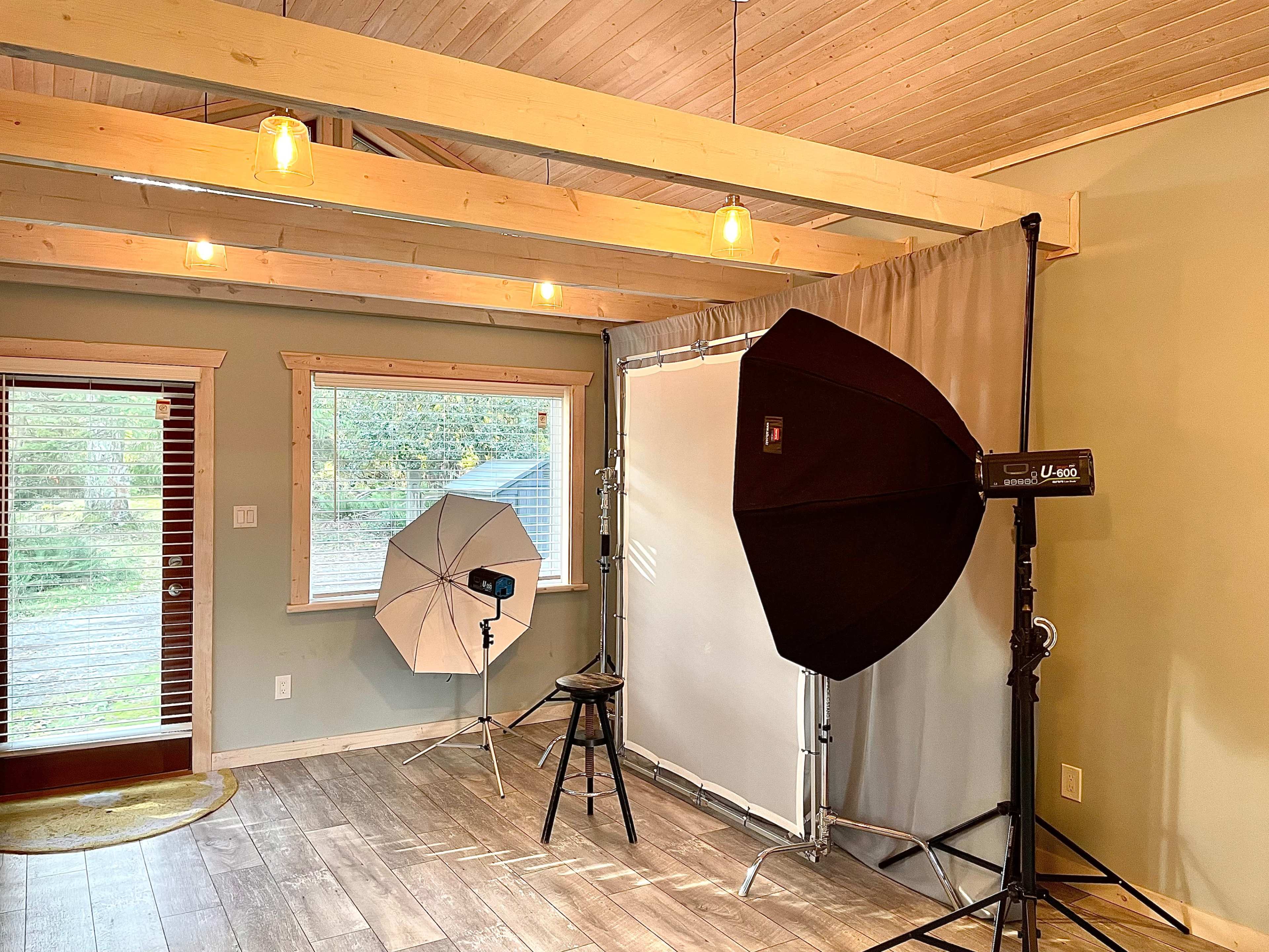 The image shows a photography studio setup featuring a backdrop, lighting equipment, and a stool in a room with wooden beams and large windows.
