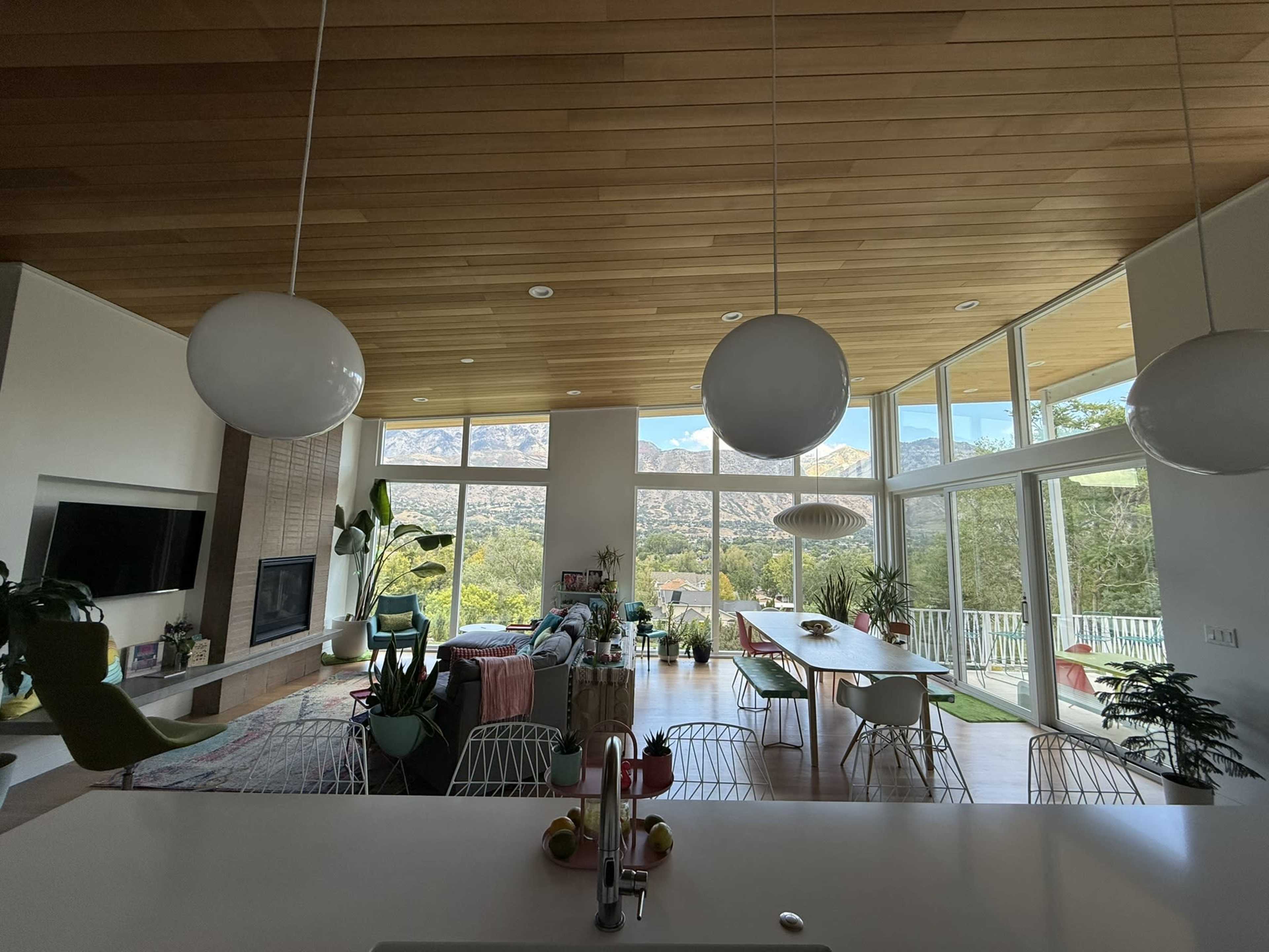 The image shows a modern living space with large windows, wooden ceilings, and a view of mountains, featuring a dining area, a lounge with plants, and pendant lighting.