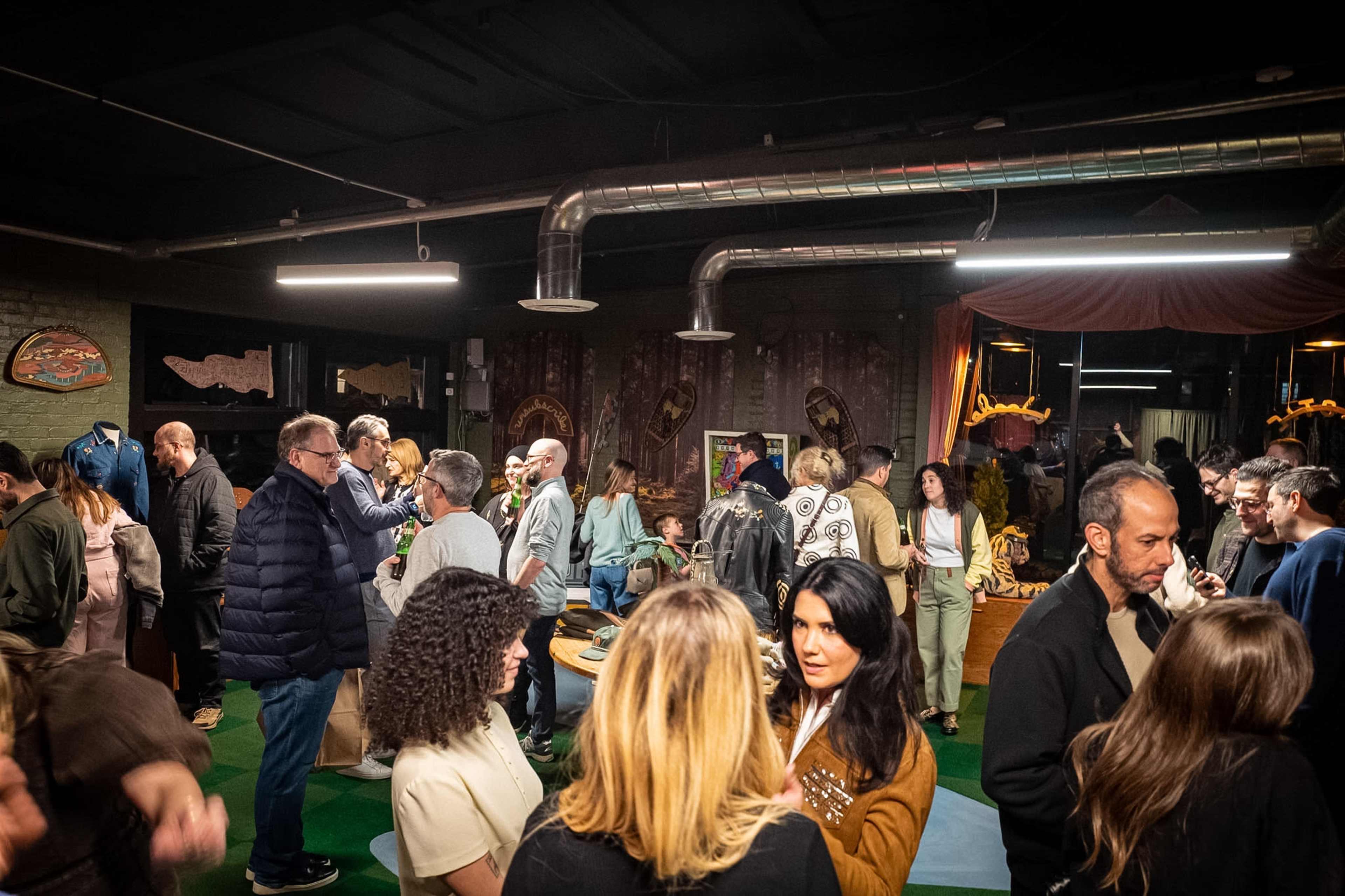 A diverse group of people congregates inside a stylish venue, engaging in conversation and browsing various displays.