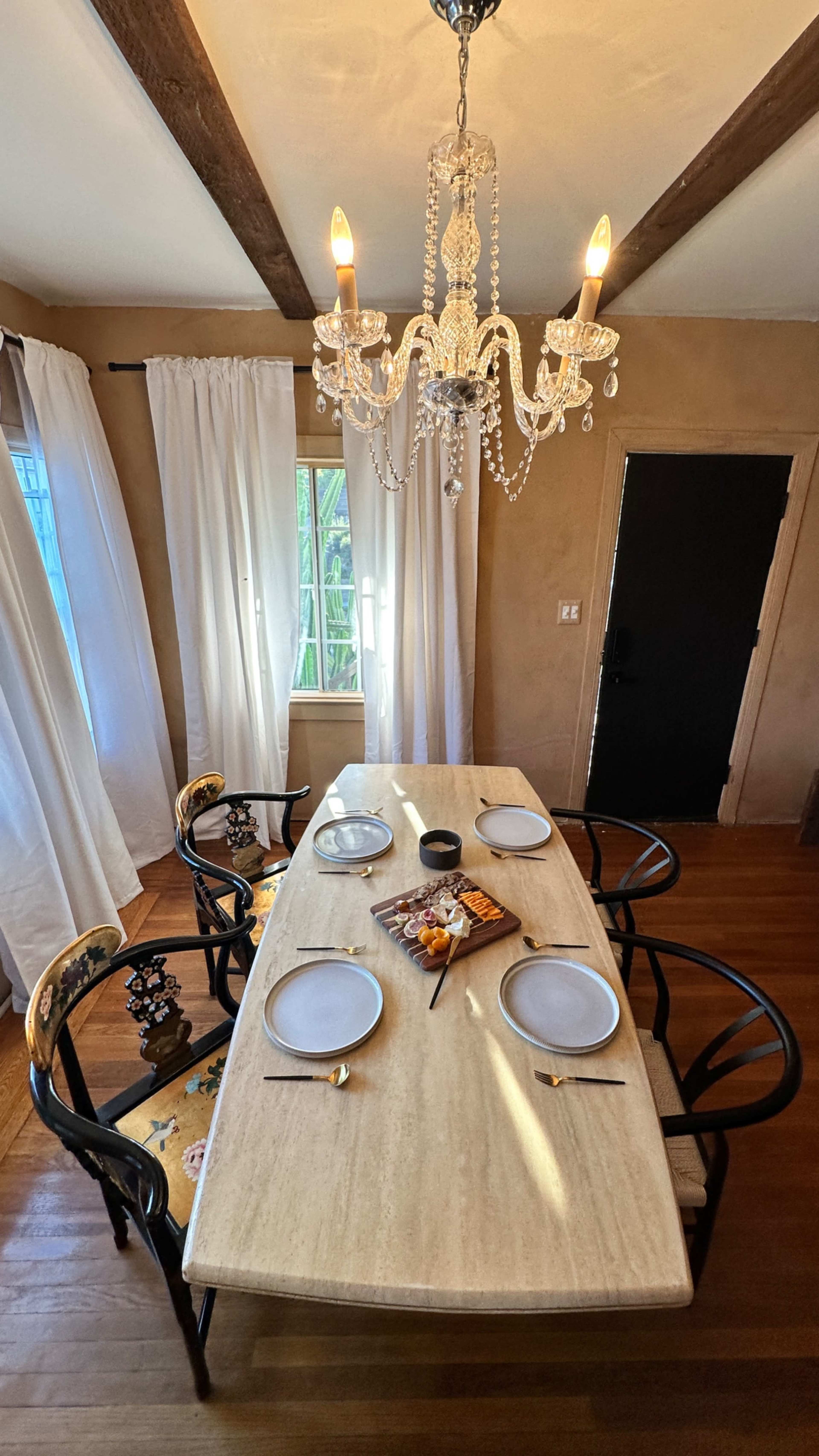A dining table is set with plates and a fruit display beneath a chandelier, surrounded by light curtains and a dark door.