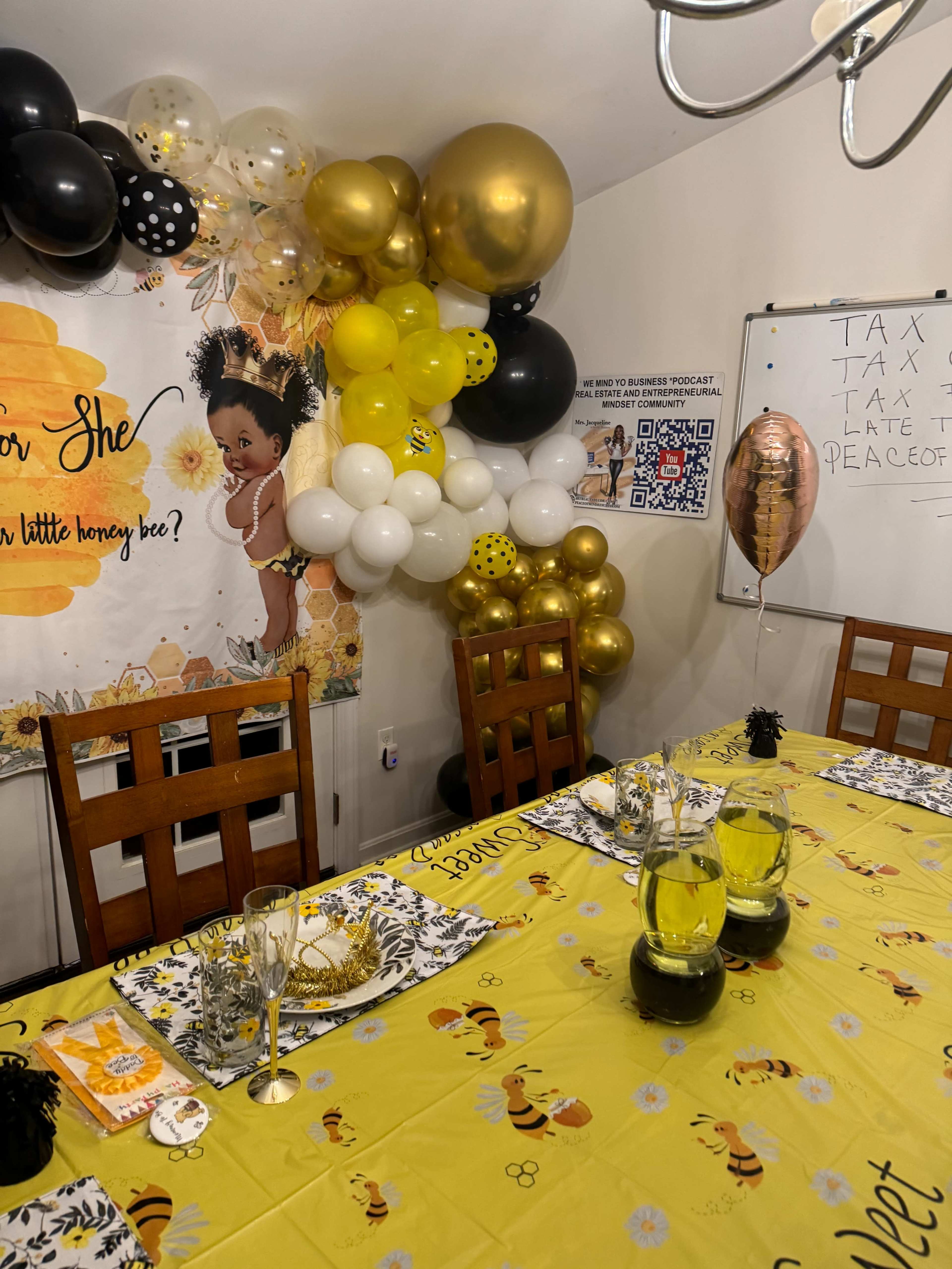 A dining table is set with yellow decorations and a backdrop featuring a bee-themed party, complete with a cluster of balloons and a whiteboard.