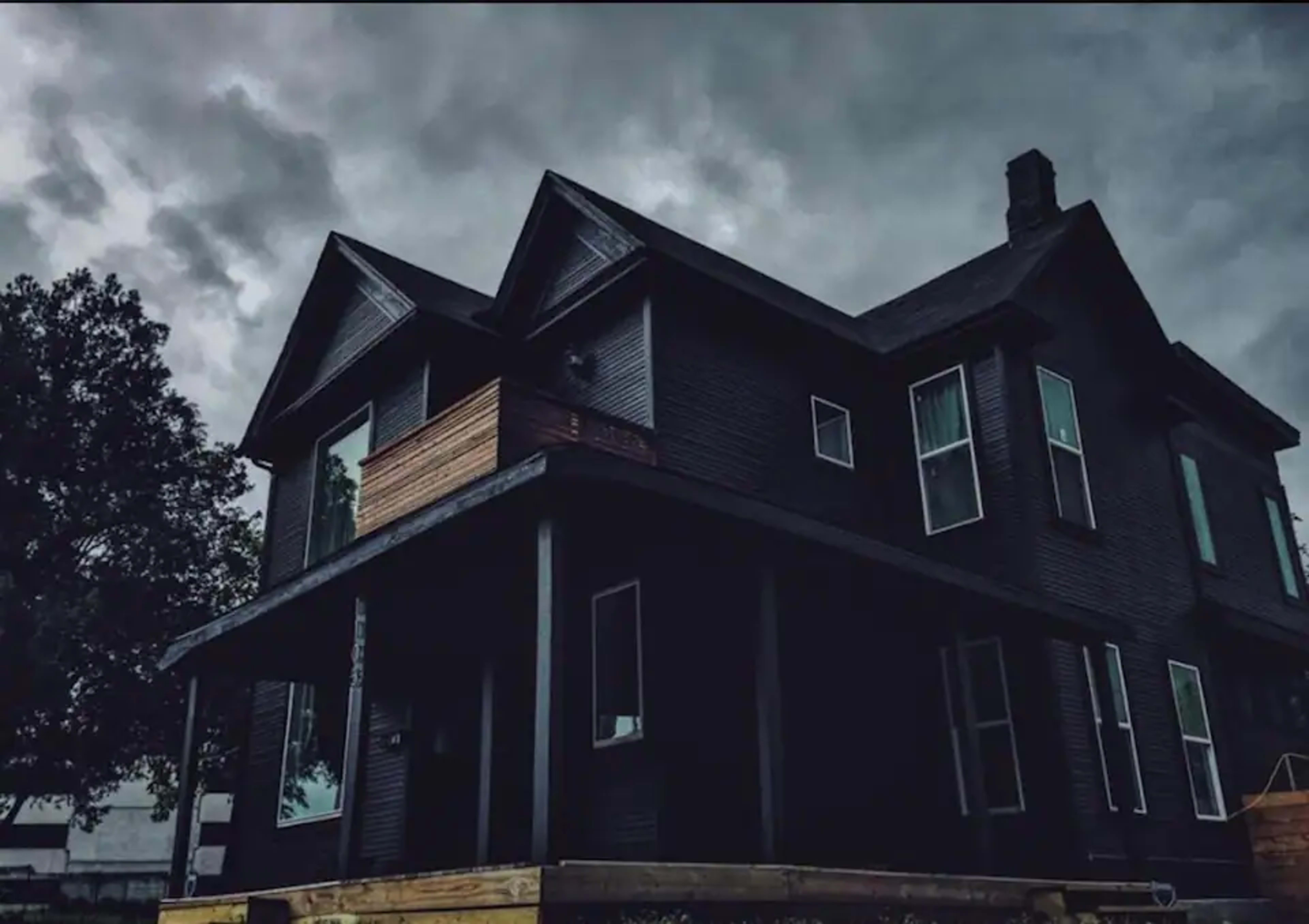 A dark, two-story house with multiple windows and a wooden balcony sits under a cloudy sky.