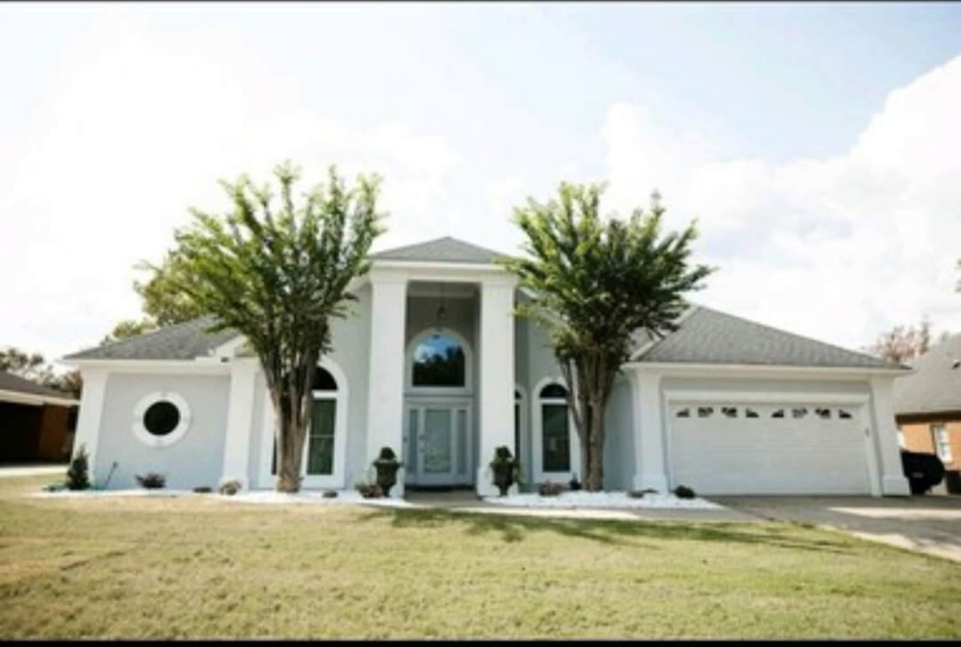 A modern, single-story house with a symmetrical facade, large front windows, and two trees flanking the entrance.