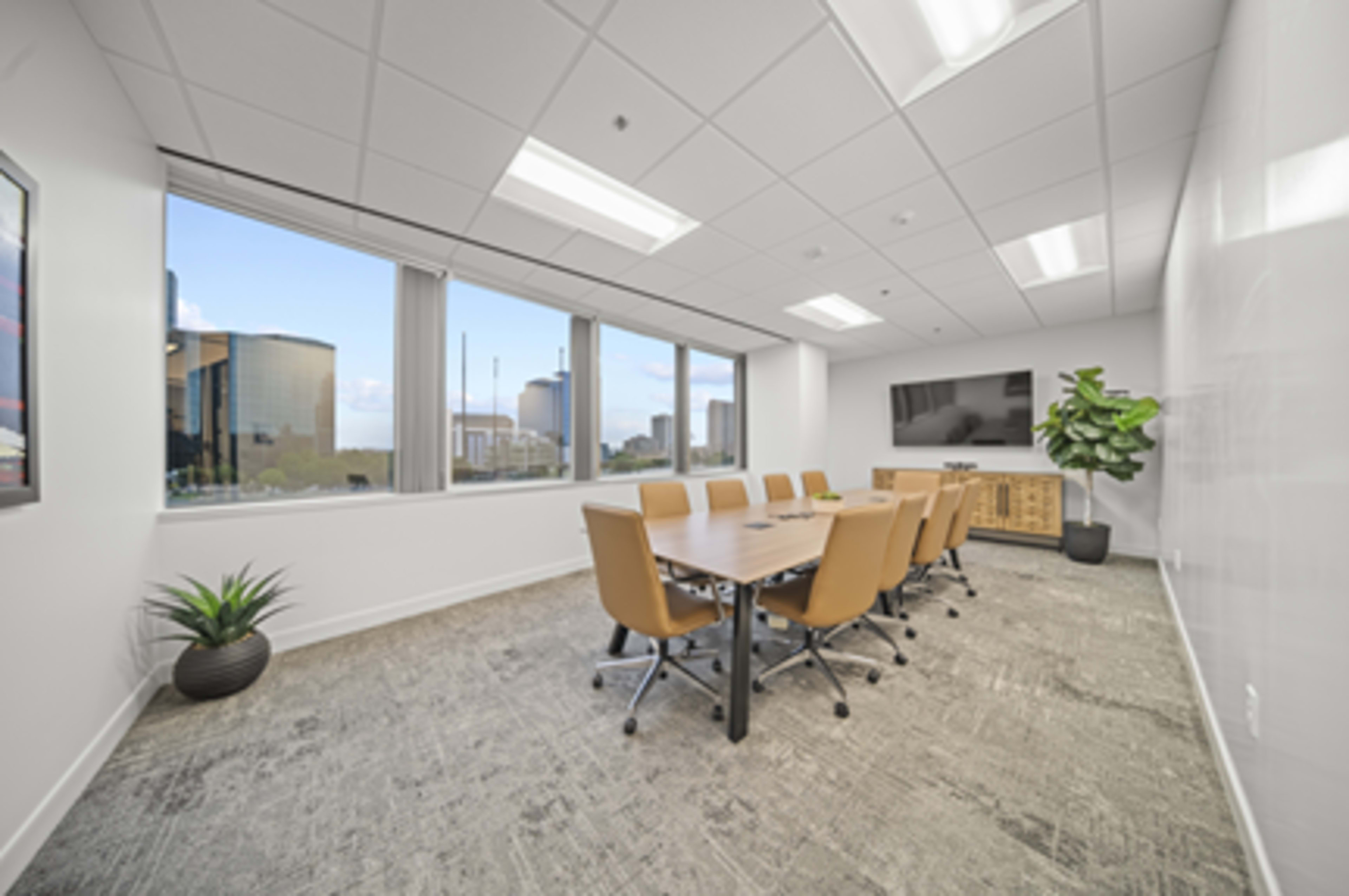 A modern conference room features a long table surrounded by ergonomic chairs, large windows, and a TV mounted on the wall.