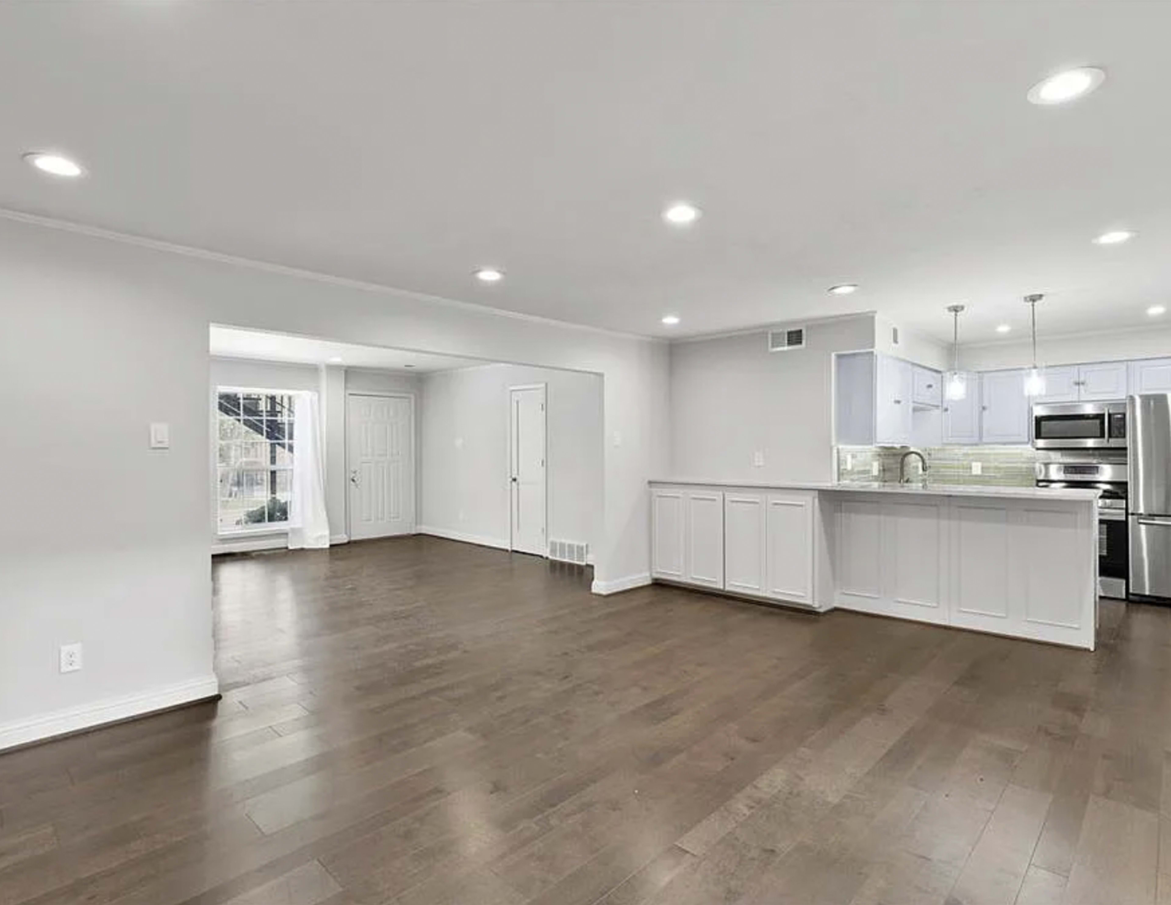 A spacious, open-concept living area with hardwood floors, white walls, and a modern kitchen featuring stainless steel appliances.