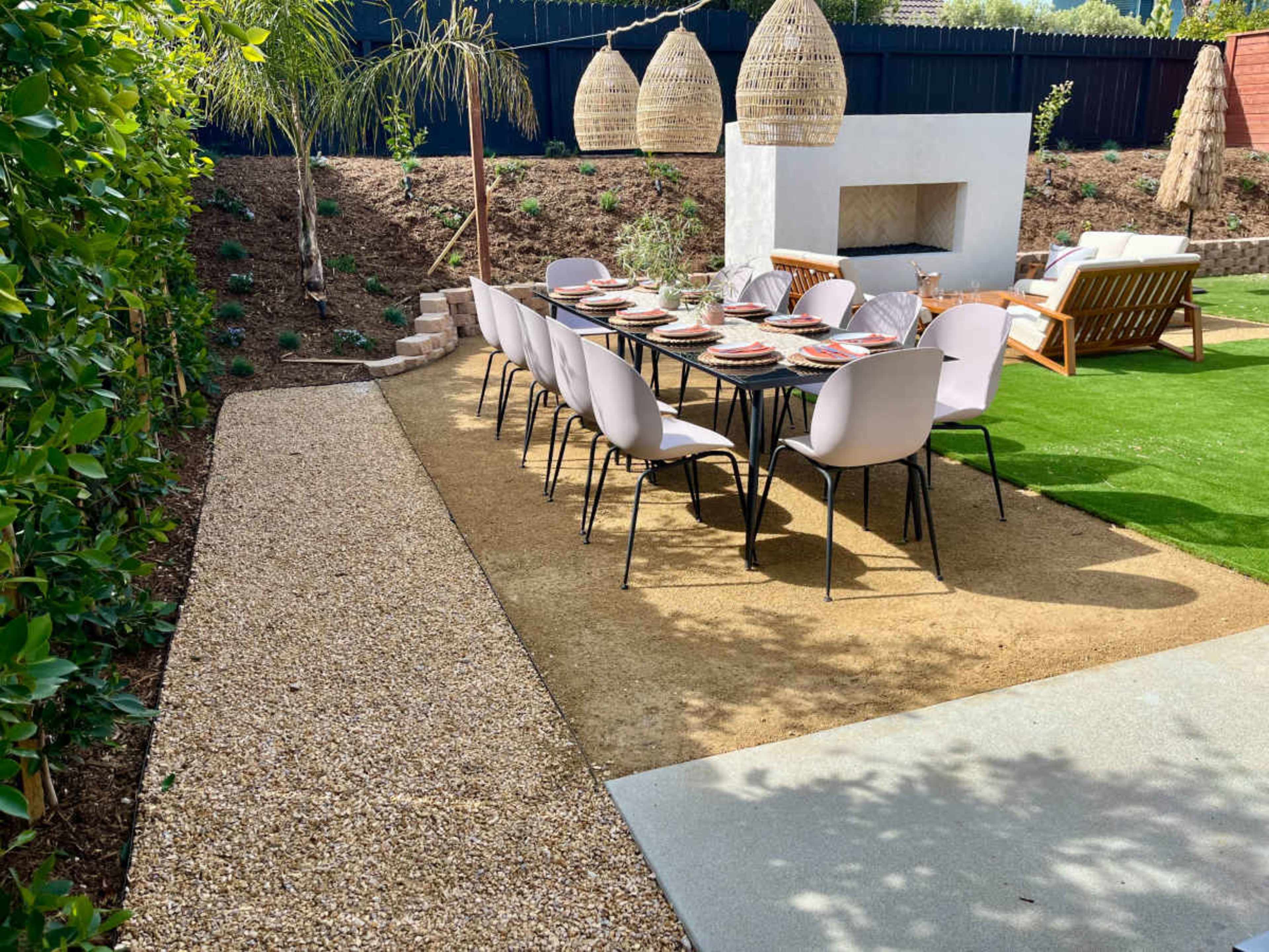 The image shows a patio area with a long dining table set for a meal, surrounded by green grass and decorative plants.