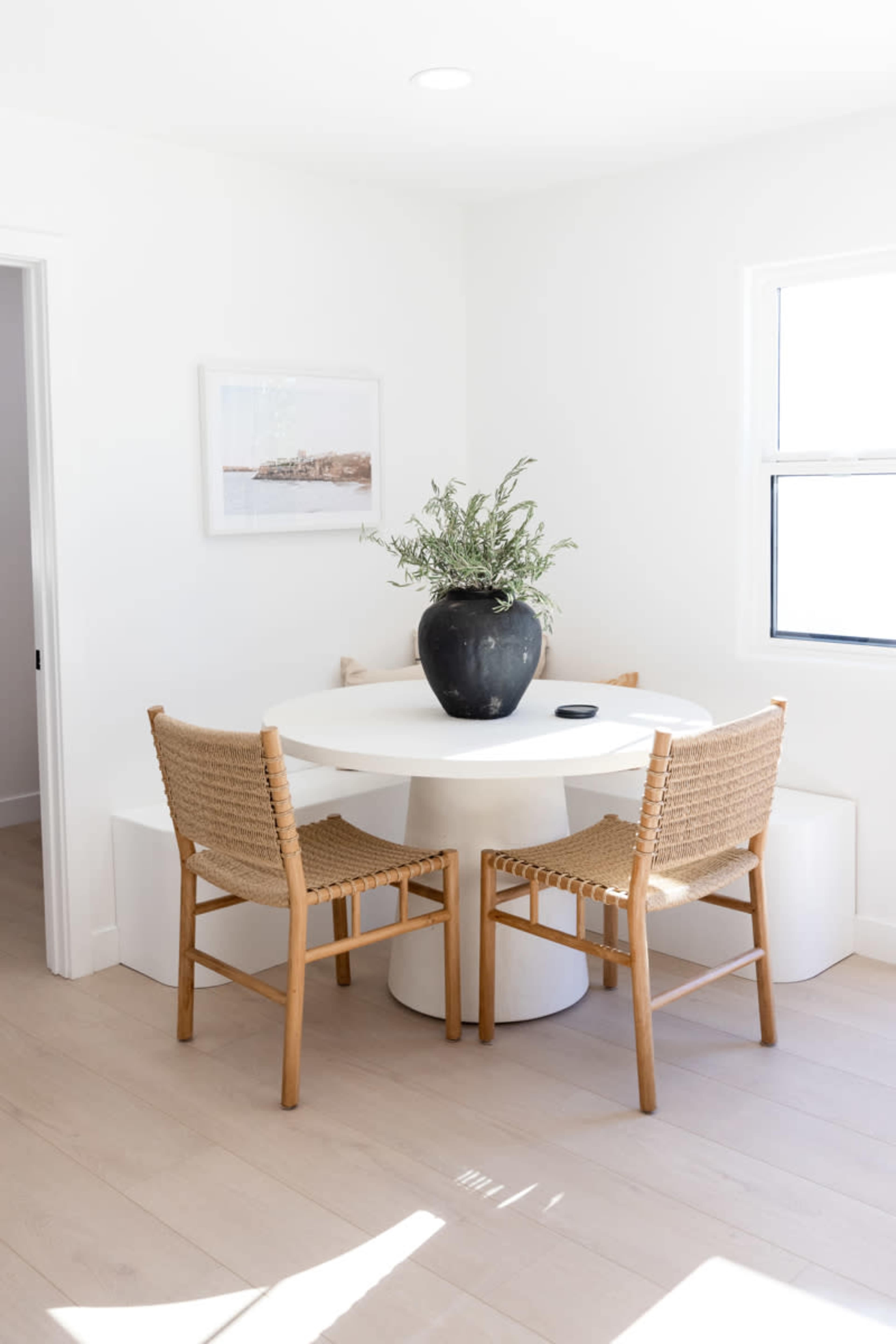 A round white dining table with two woven chairs and a large black vase filled with plants is positioned in a bright, minimalist room.