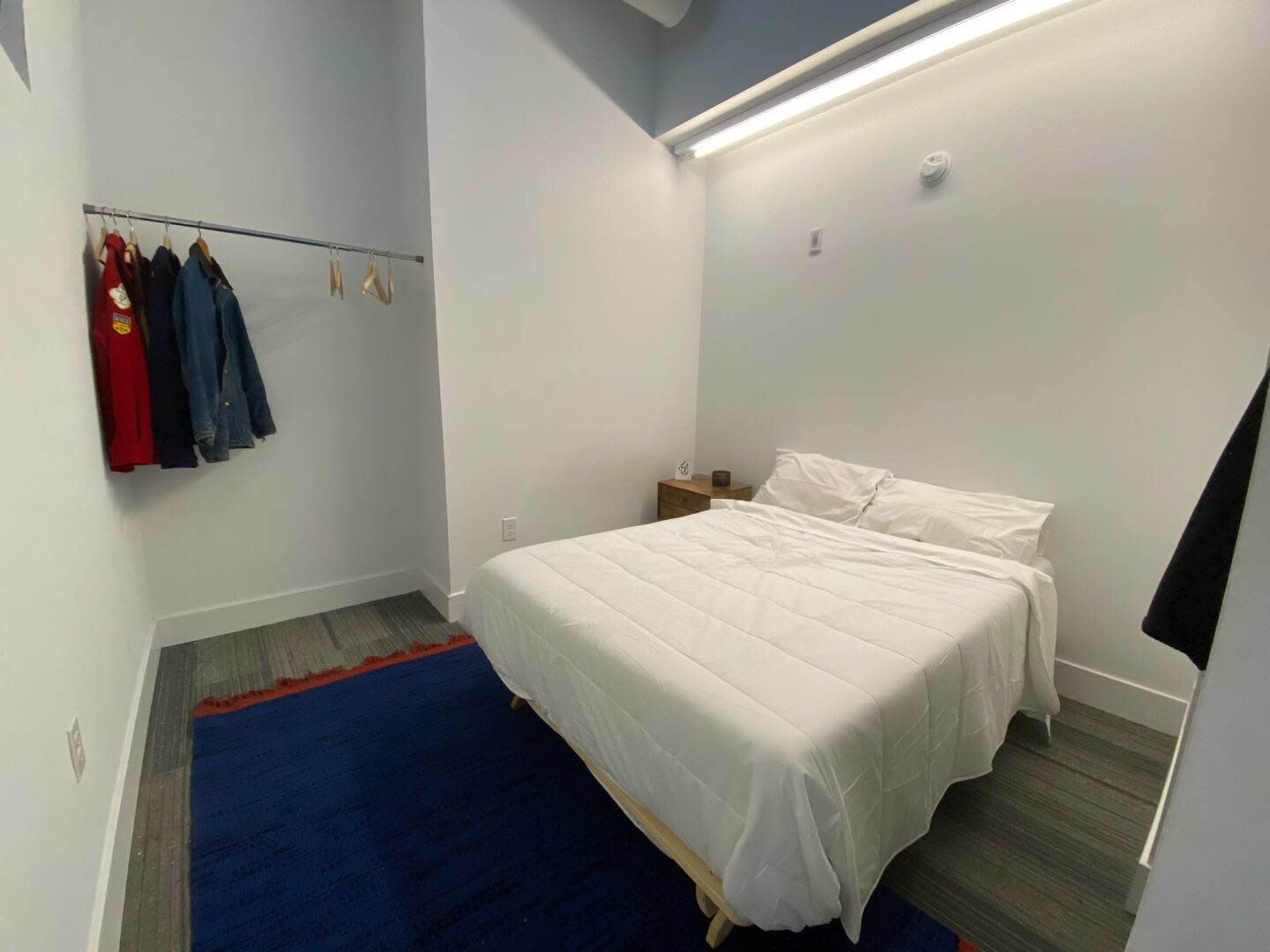A small bedroom with a white bed, a hanging clothes rack on the left, and a wooden nightstand beside the bed.