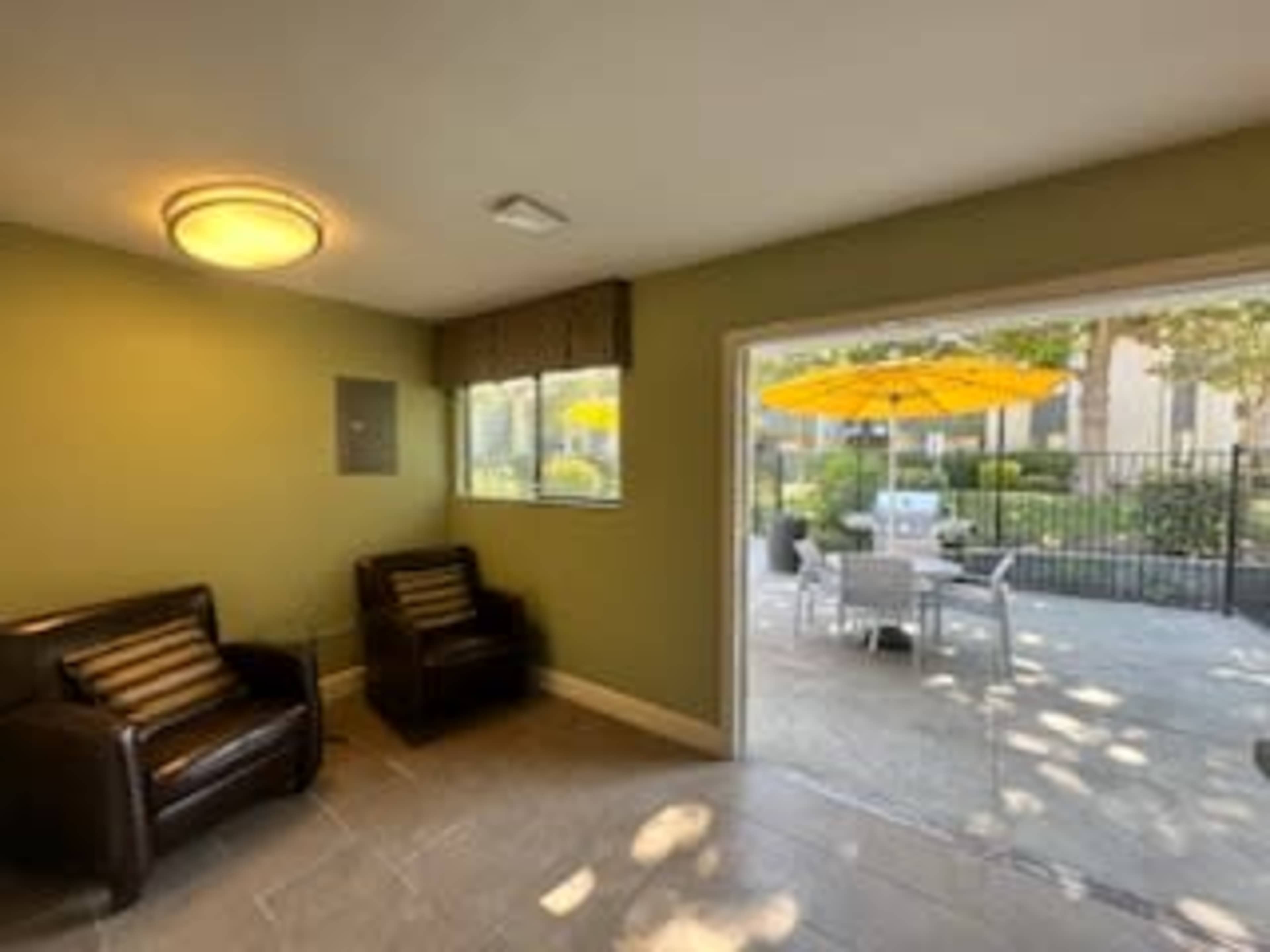 A small indoor area with two black armchairs, a window looking out to a patio with an umbrella, and a table with chairs visible in the background.