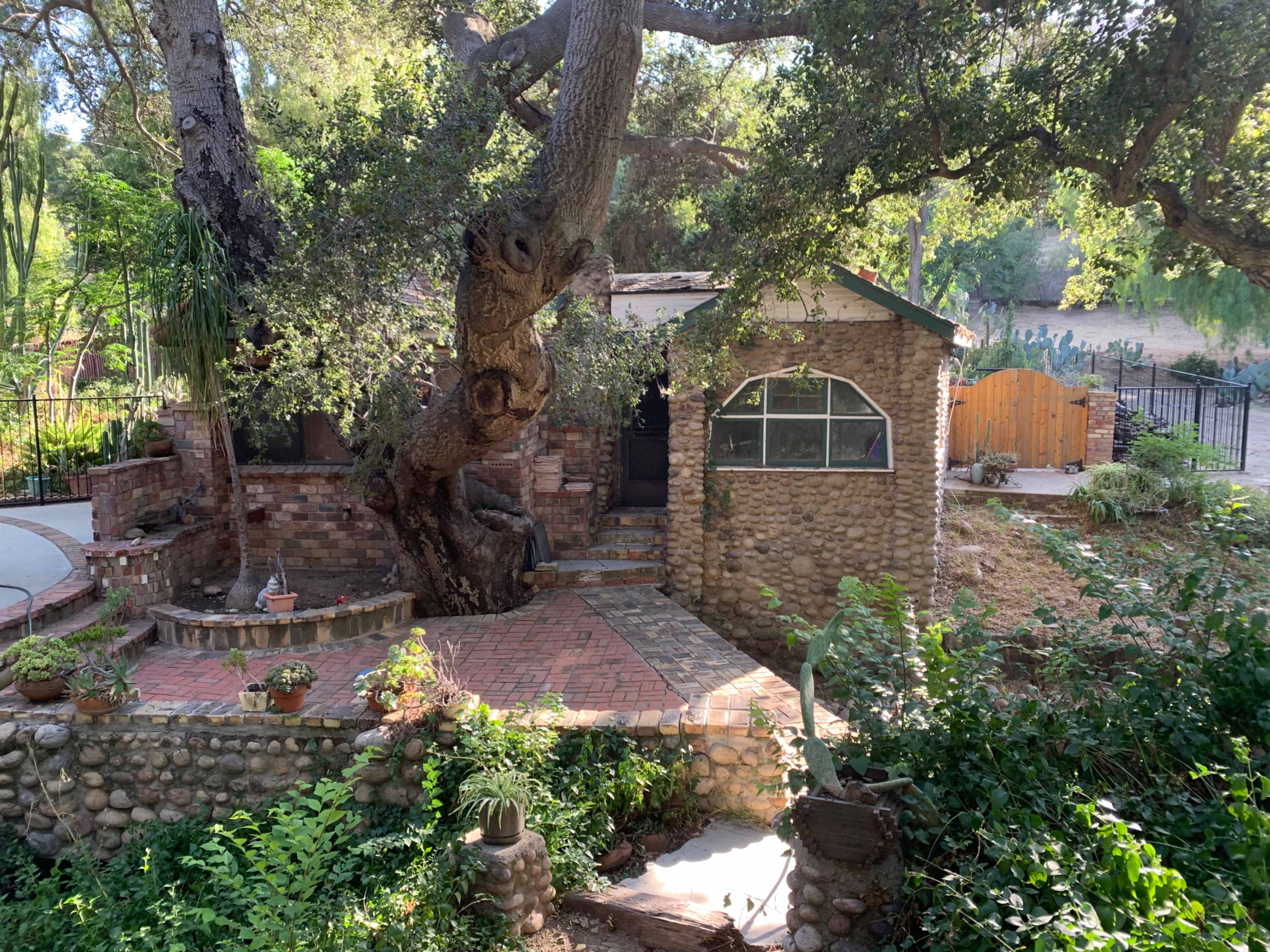 A rustic stone house with a large tree at its side and a winding brick pathway leads toward the entrance, surrounded by greenery.