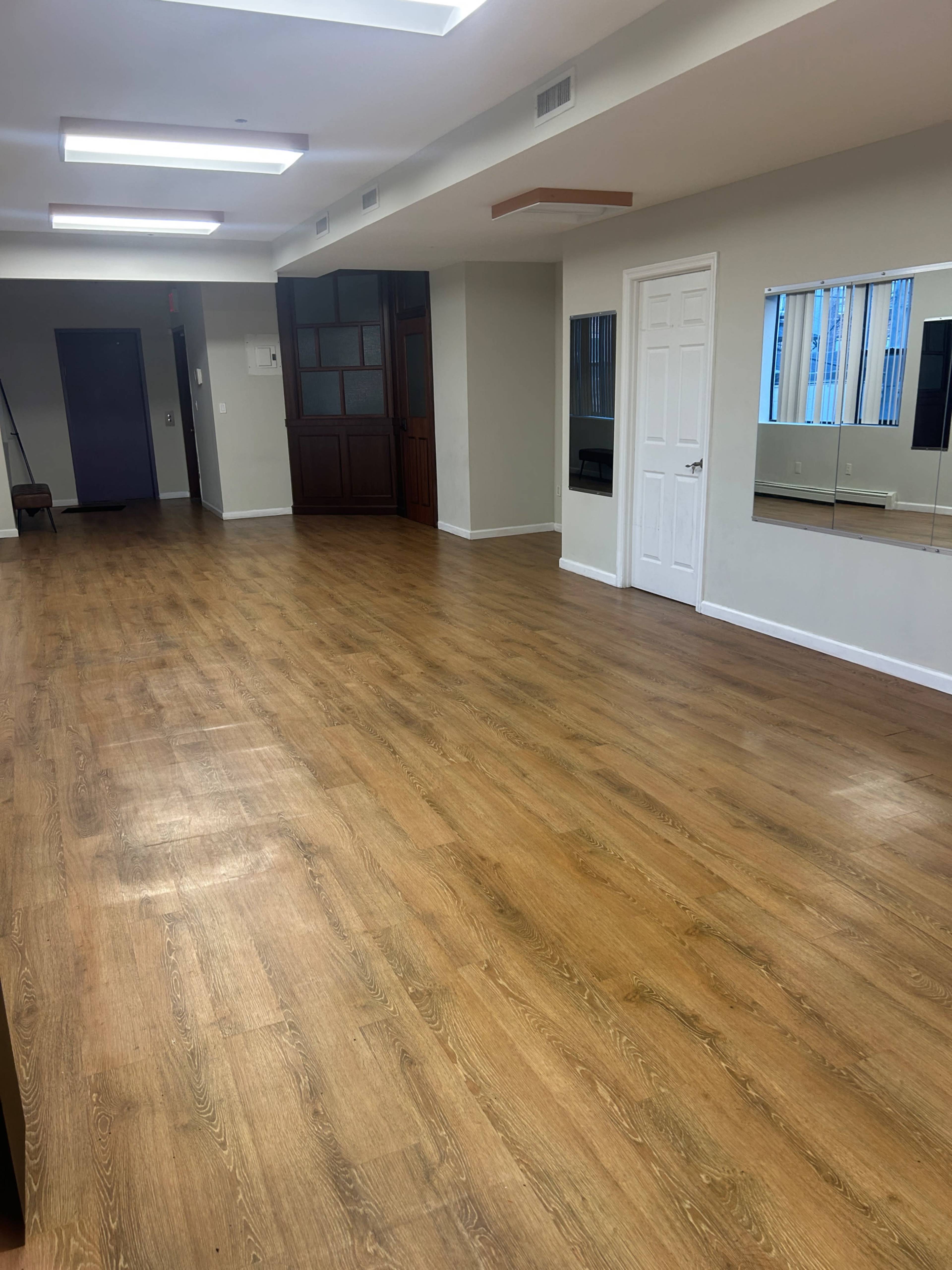 The image shows a spacious room with wooden flooring, white walls, and a few mirrors mounted on one wall.