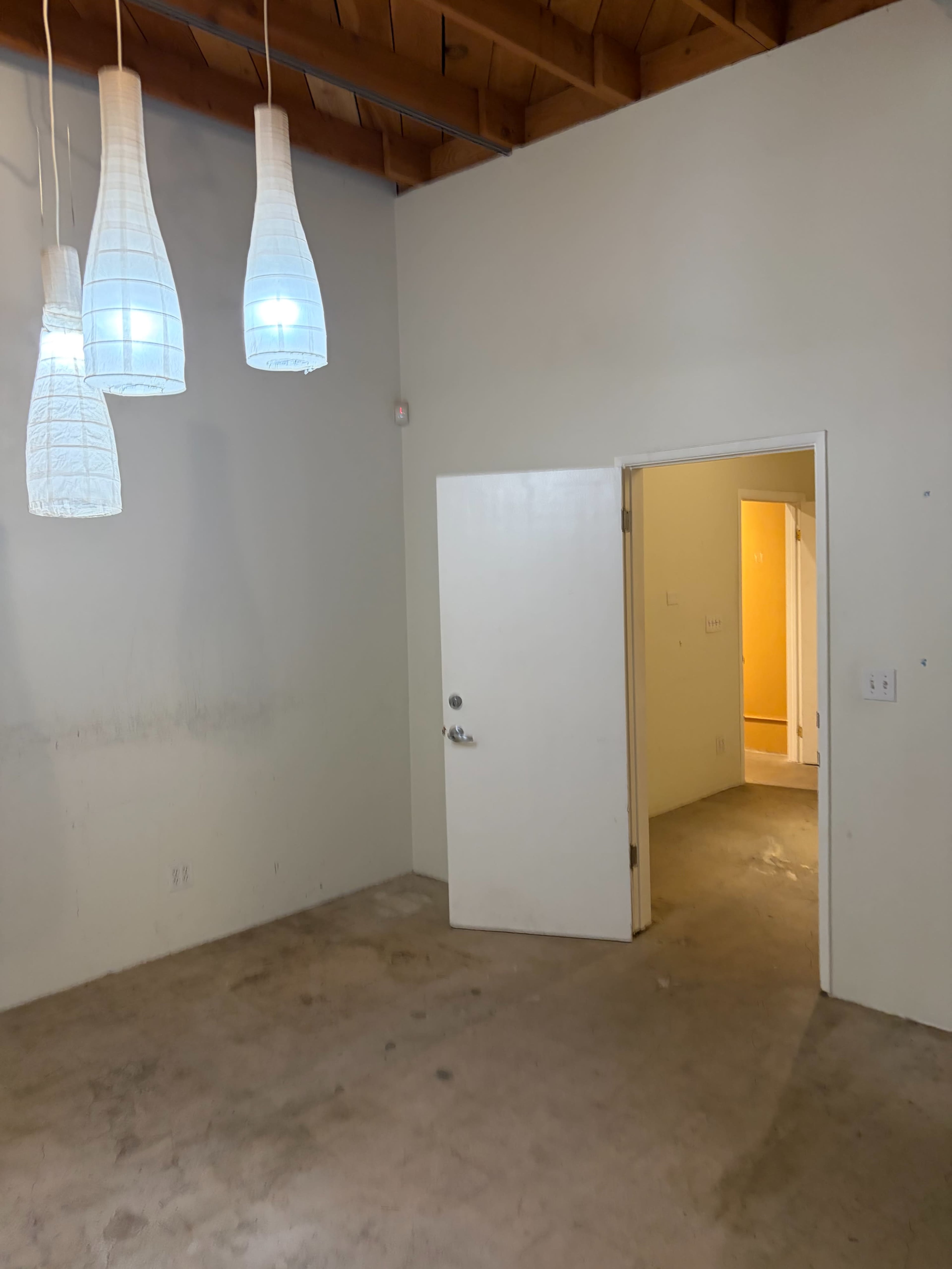 The image shows a spacious, empty room with three hanging light fixtures and an open door leading to another area with a yellow wall visible in the background.