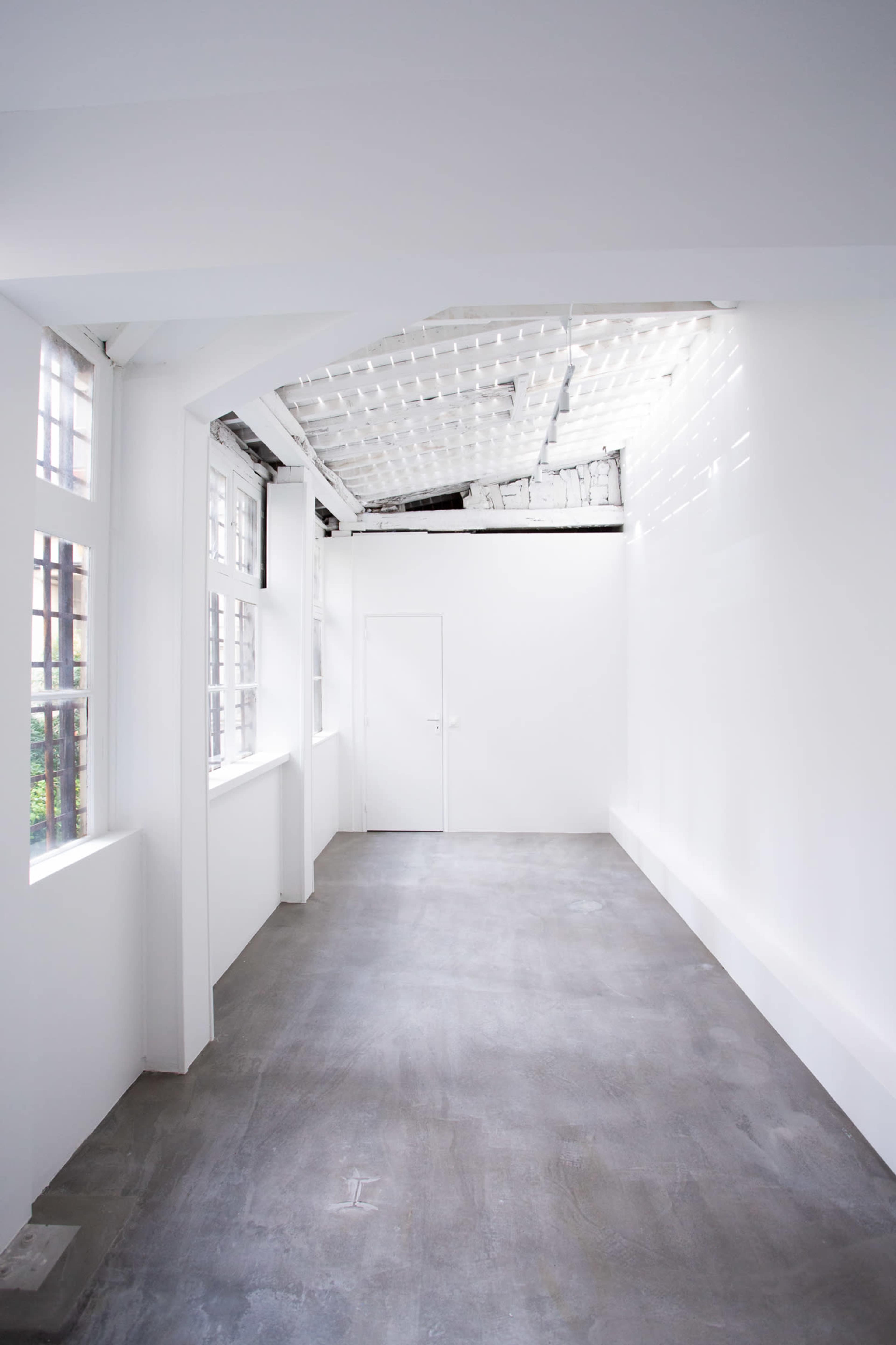 The image shows a minimalist white room with high ceilings, large windows, and a smooth concrete floor.