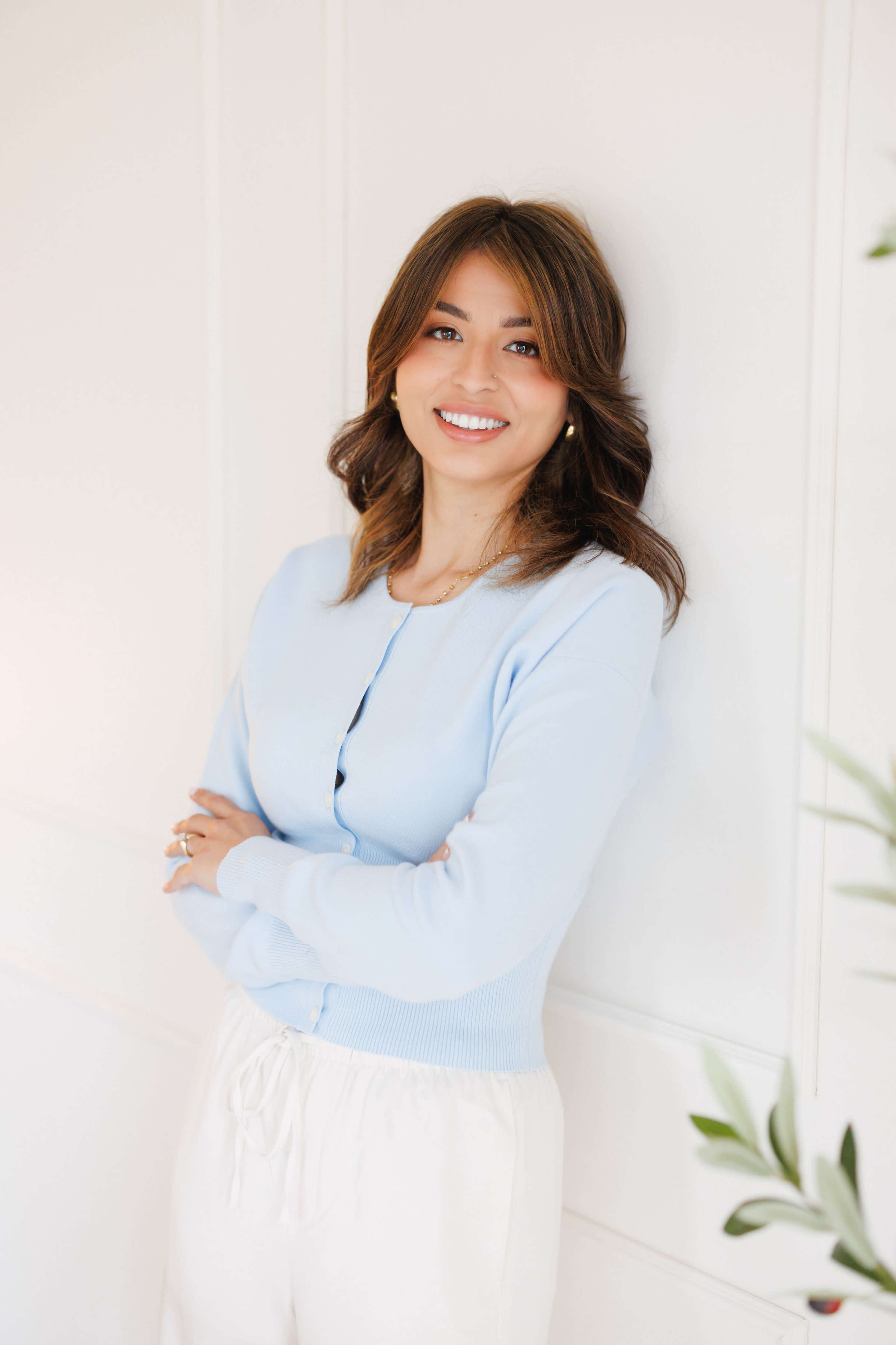 A woman with shoulder-length brown hair, wearing a light blue cardigan and white pants, stands smiling with her arms crossed against a white wall.