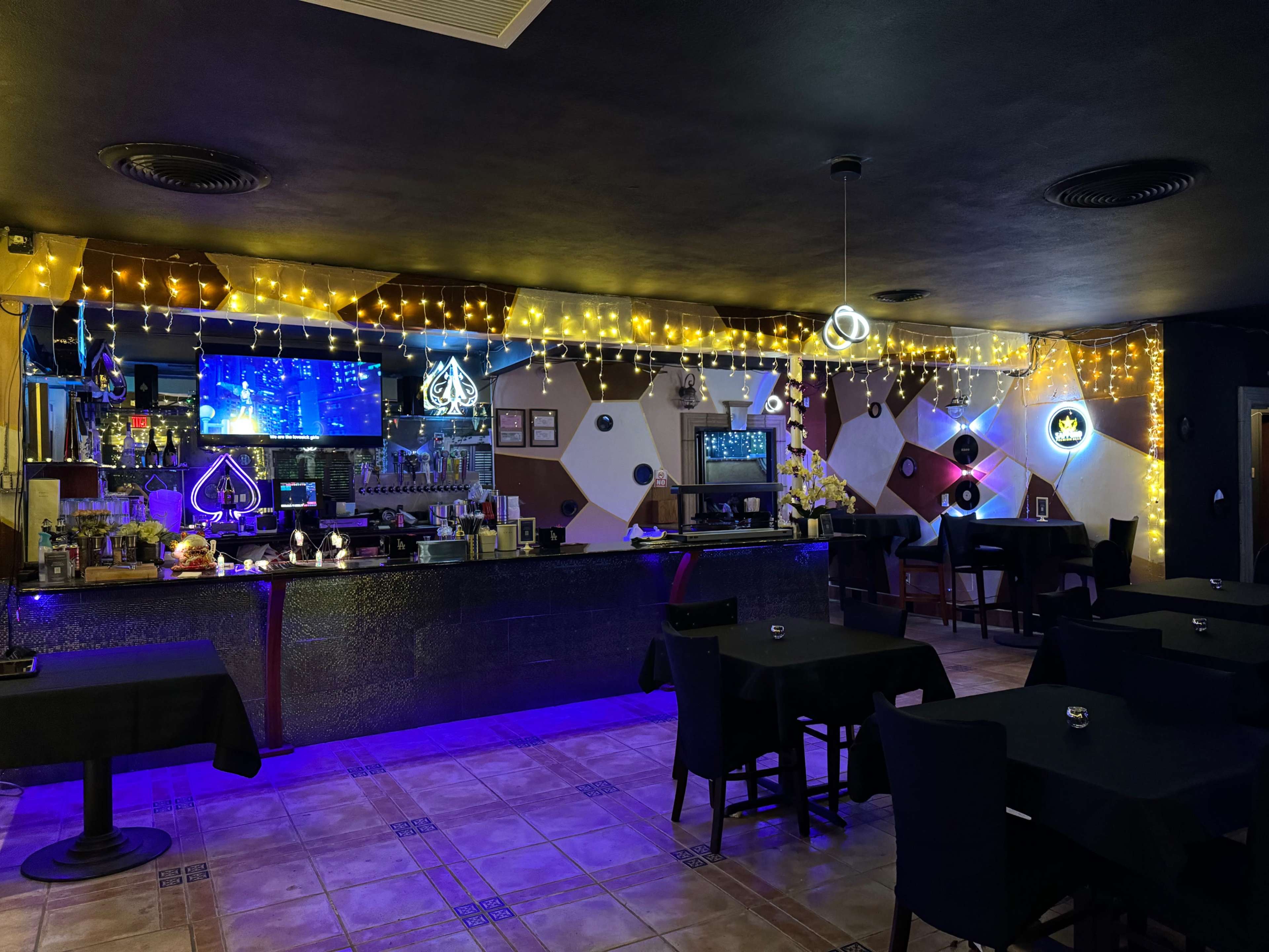 A dimly lit bar area with black tables, a long bar counter adorned with fairy lights, and multiple screens displaying graphics in the background.