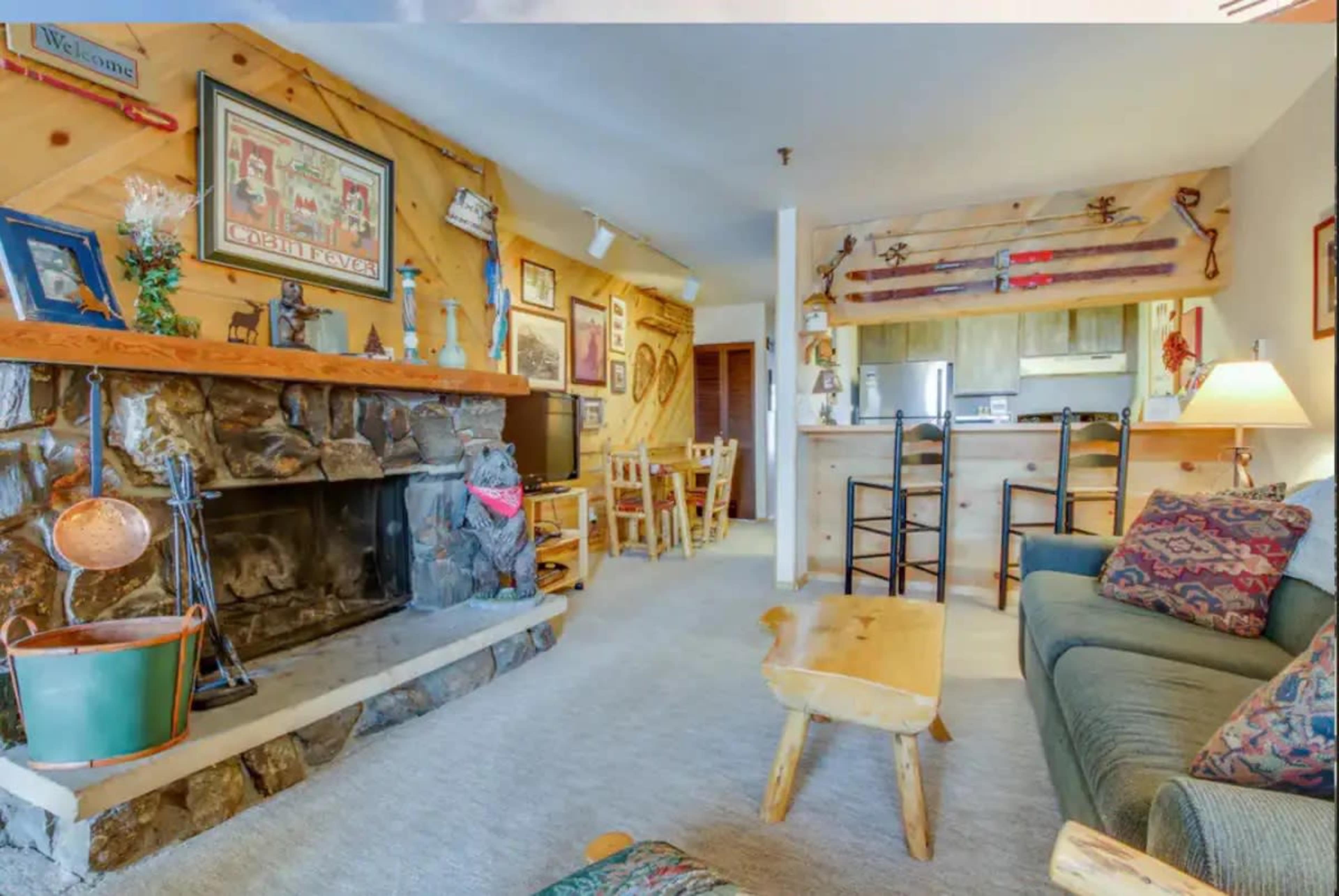 The image shows a cozy living room with a stone fireplace, wooden accents, and a kitchen in the background.