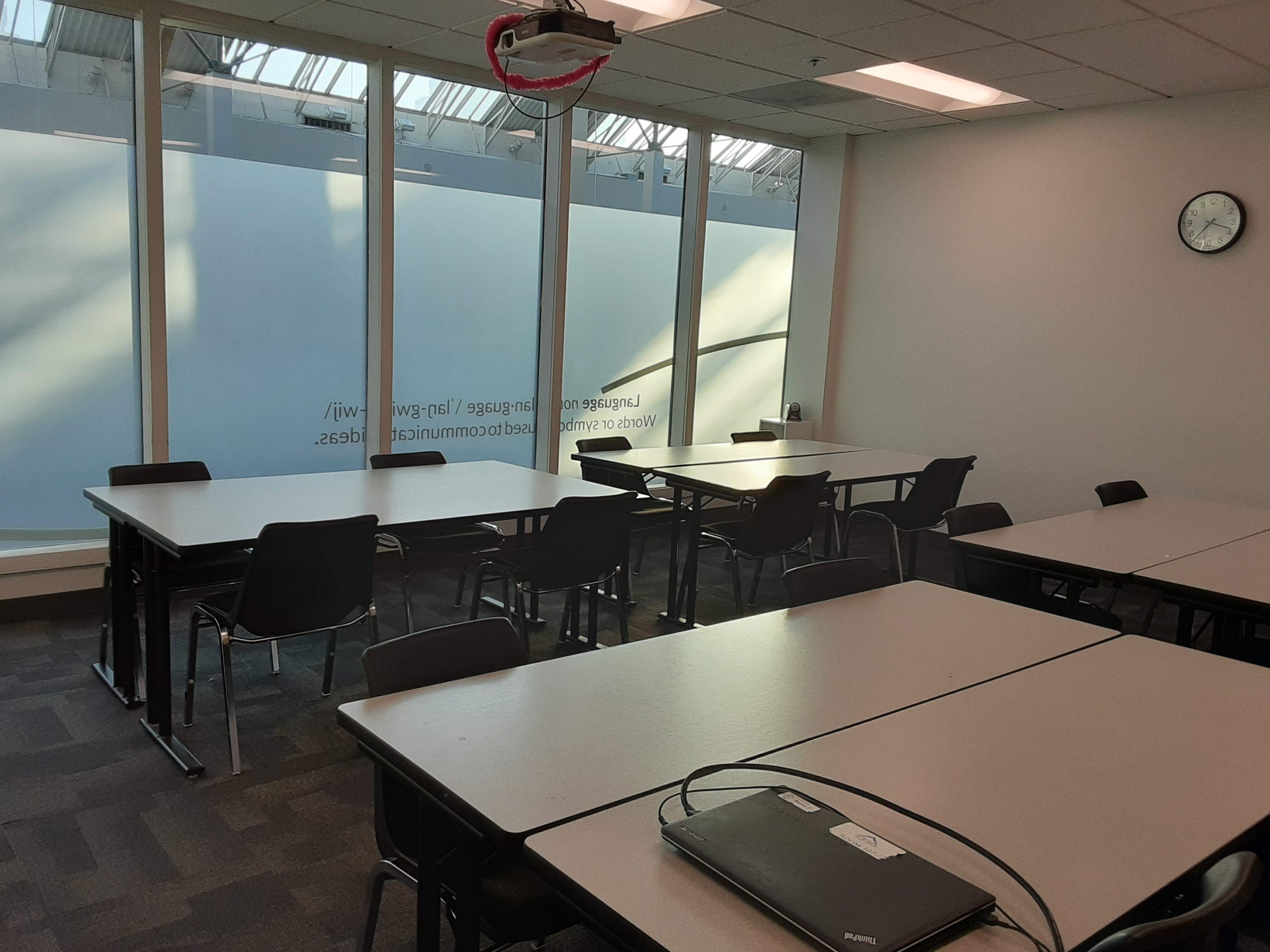 The image shows a well-lit classroom with multiple tables and chairs arranged in two groups, featuring large windows on one side.