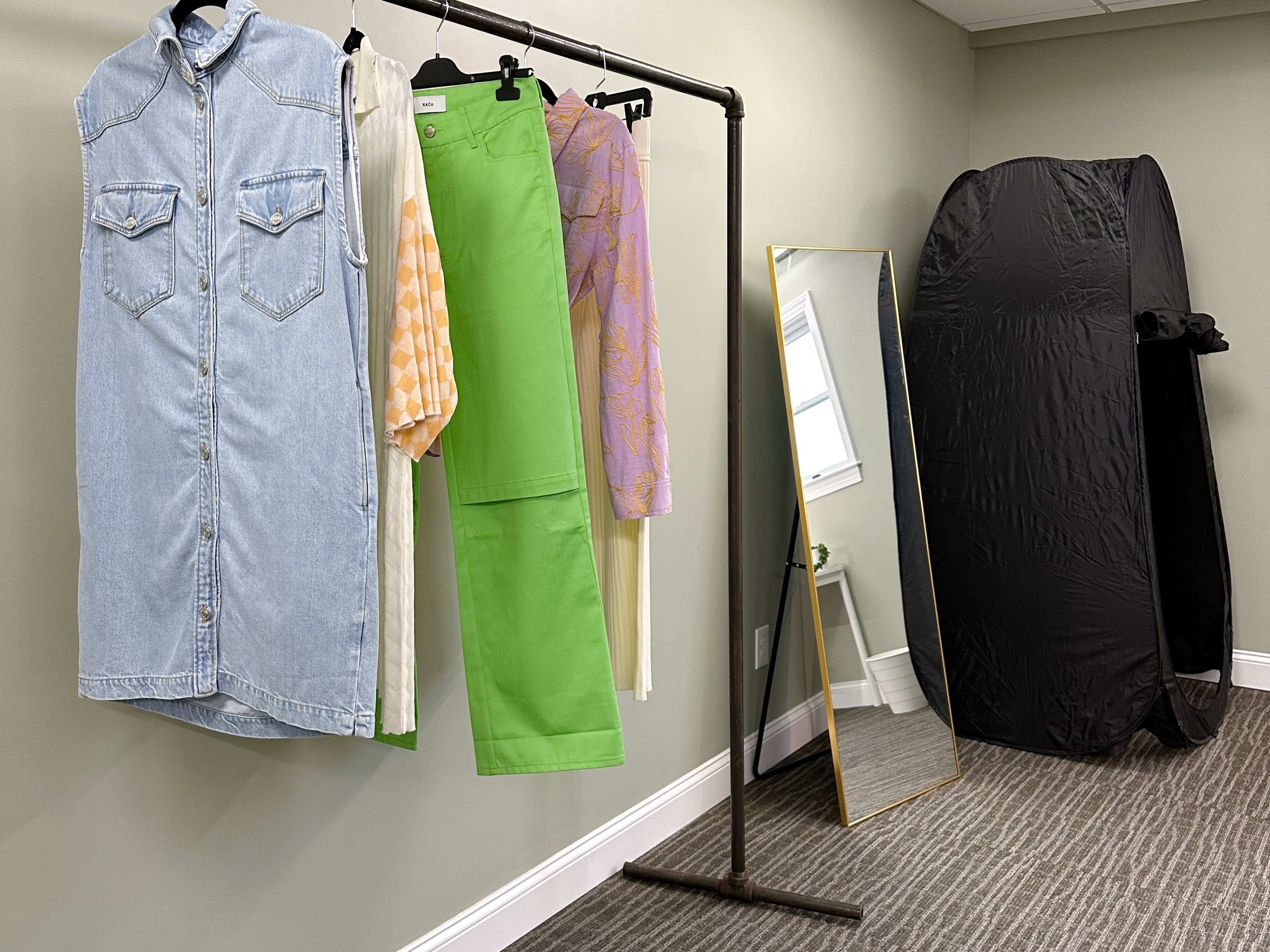 A clothing rack with various garments hanging on it, alongside a large mirror and a black clothing backdrop in a green-walled room.