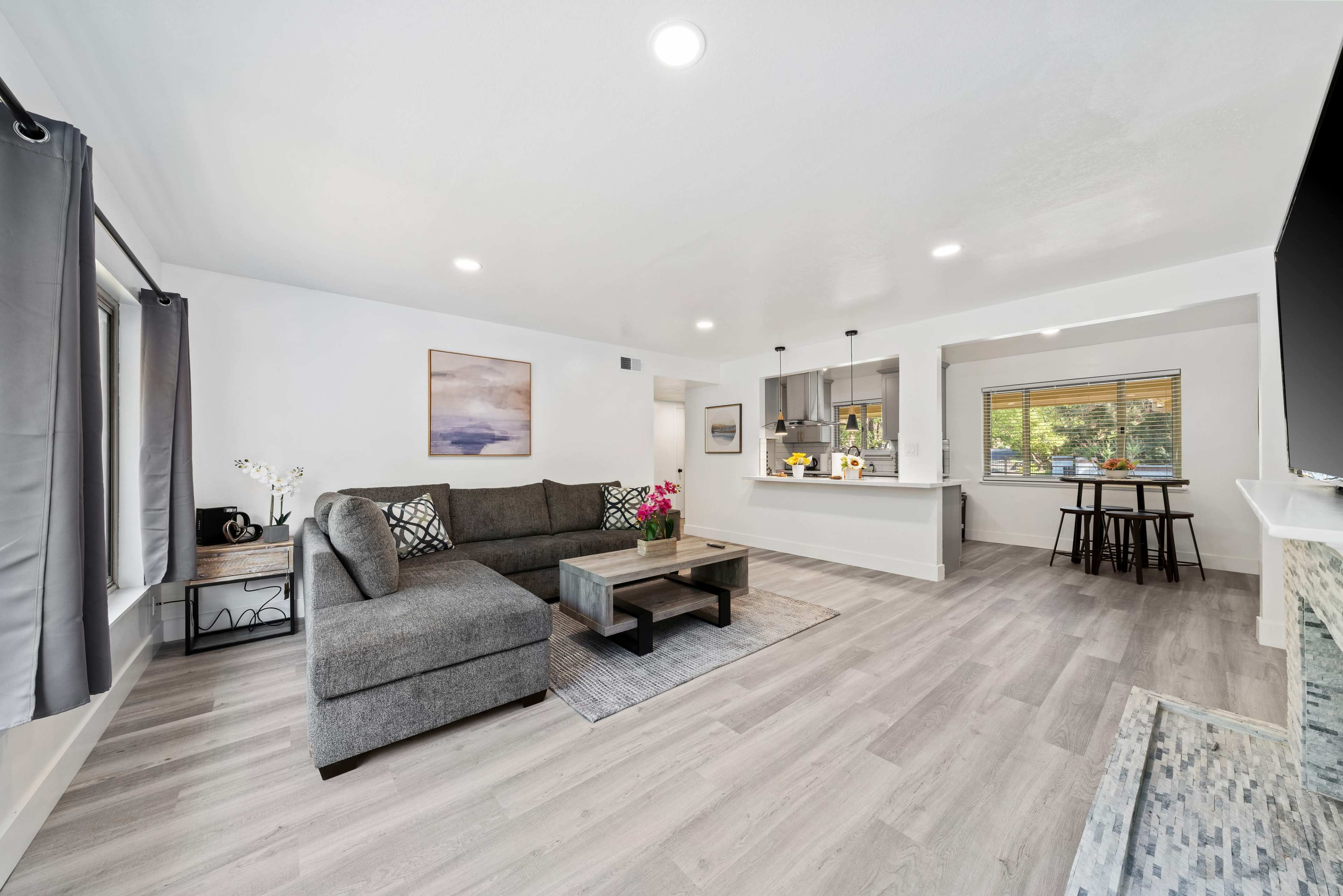The spacious living room features a gray sectional sofa, a coffee table, and a view into a modern kitchen with bar seating and natural light.