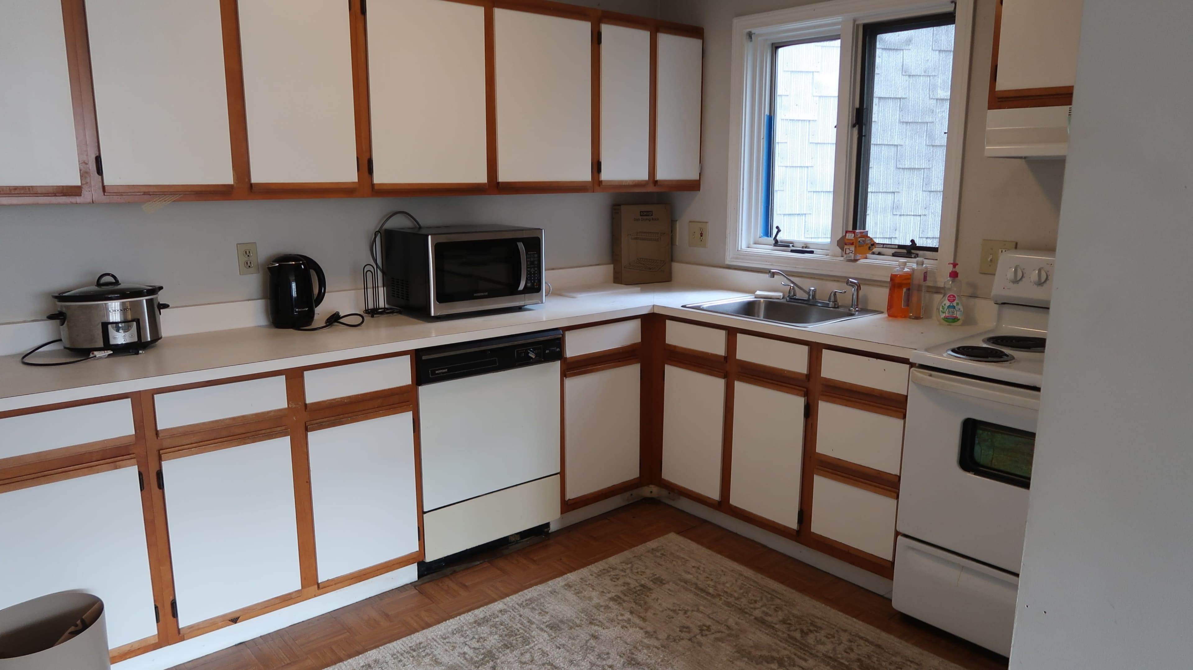 The kitchen features white cabinetry with wooden trim, a microwave, a sink, and a stove, all arranged along the walls with a window providing natural light.