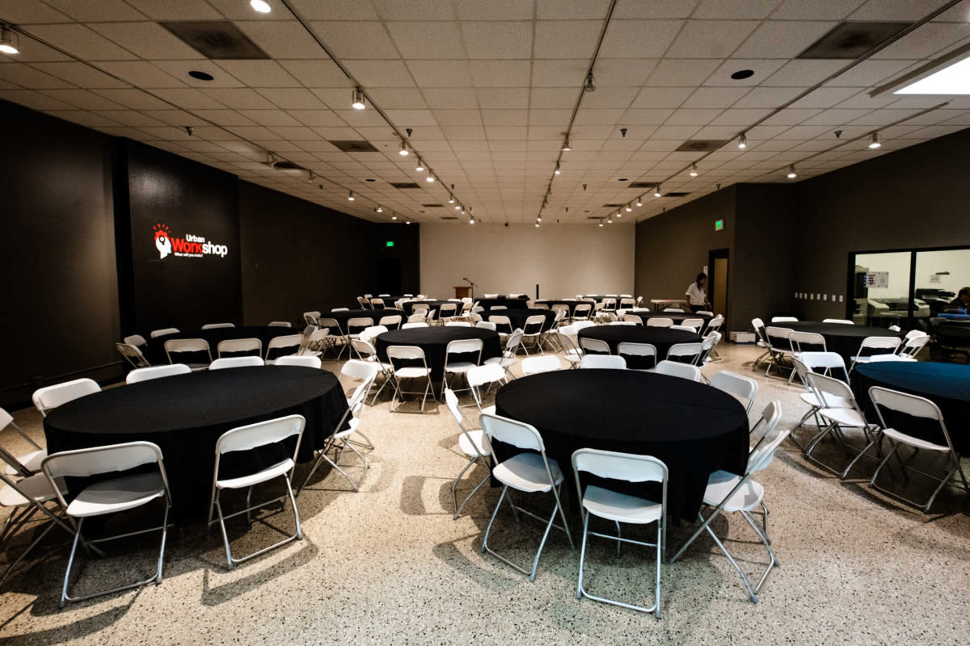 A large meeting room is arranged with multiple round tables covered in black cloth and surrounded by white chairs.