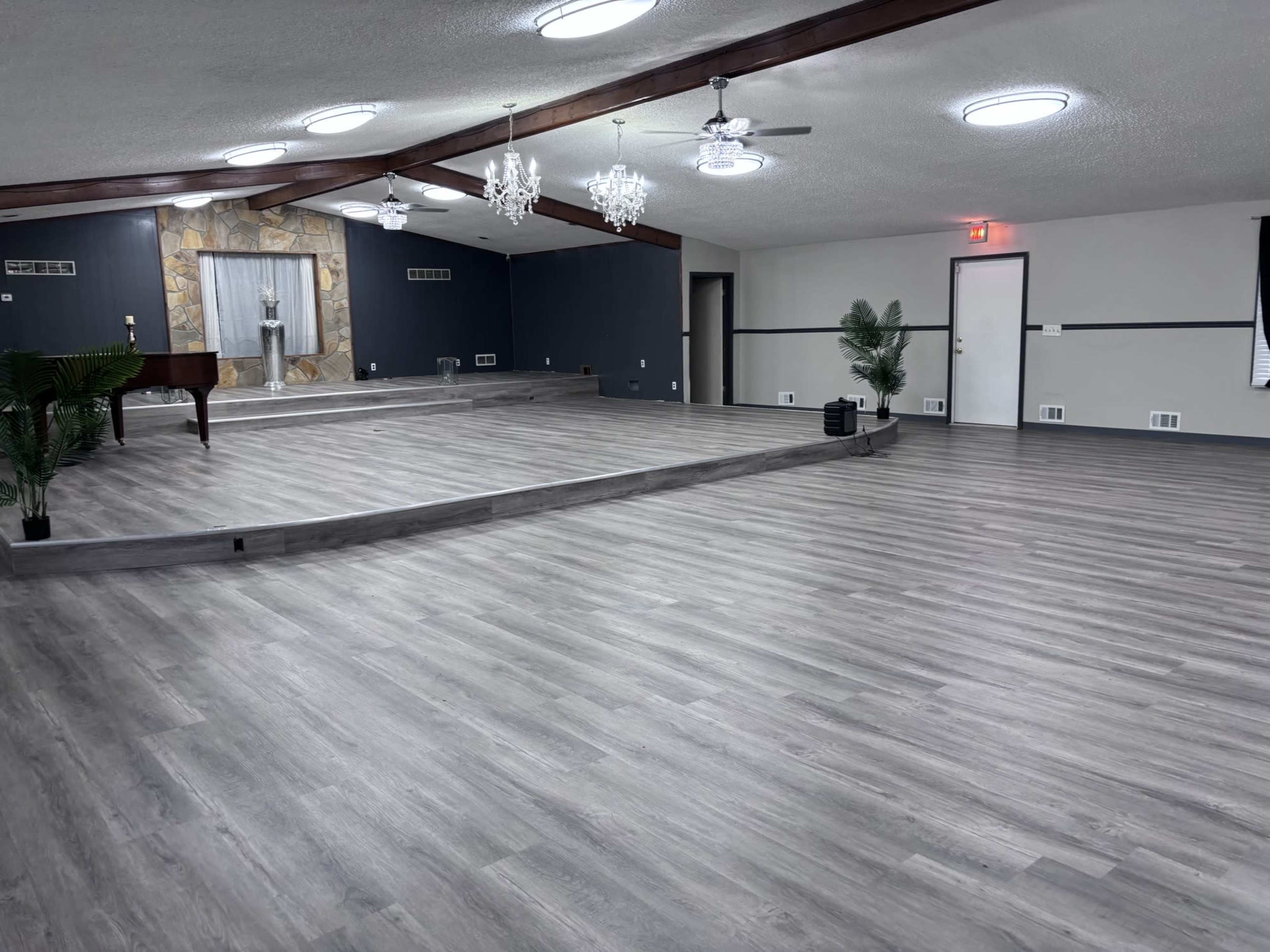 The image shows a spacious hall with wooden beams, light-colored flooring, and a small stage area at one end.