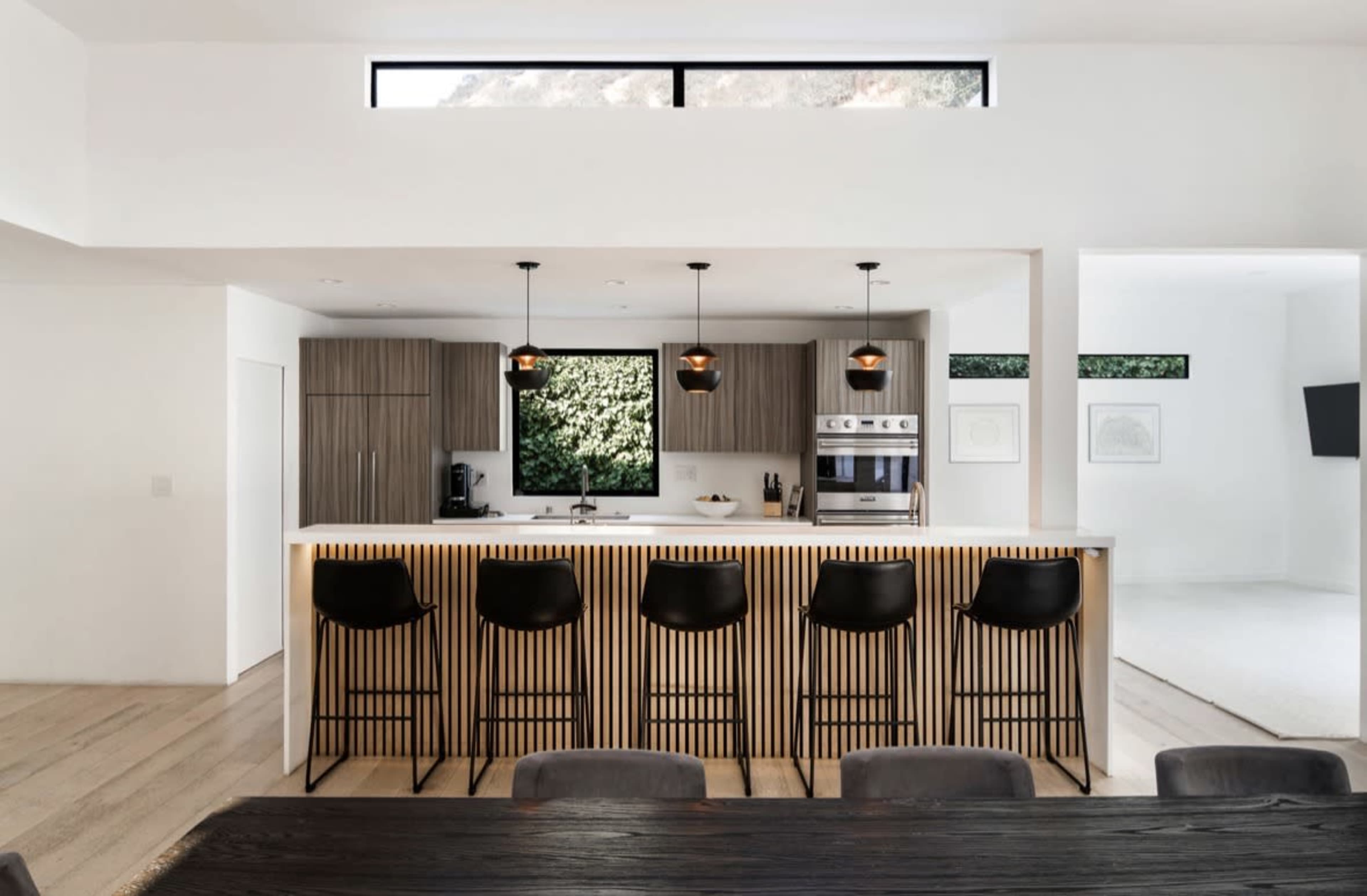 The image depicts a modern kitchen with a breakfast bar that includes black chairs, wooden cabinetry, and large windows allowing natural light to enter.