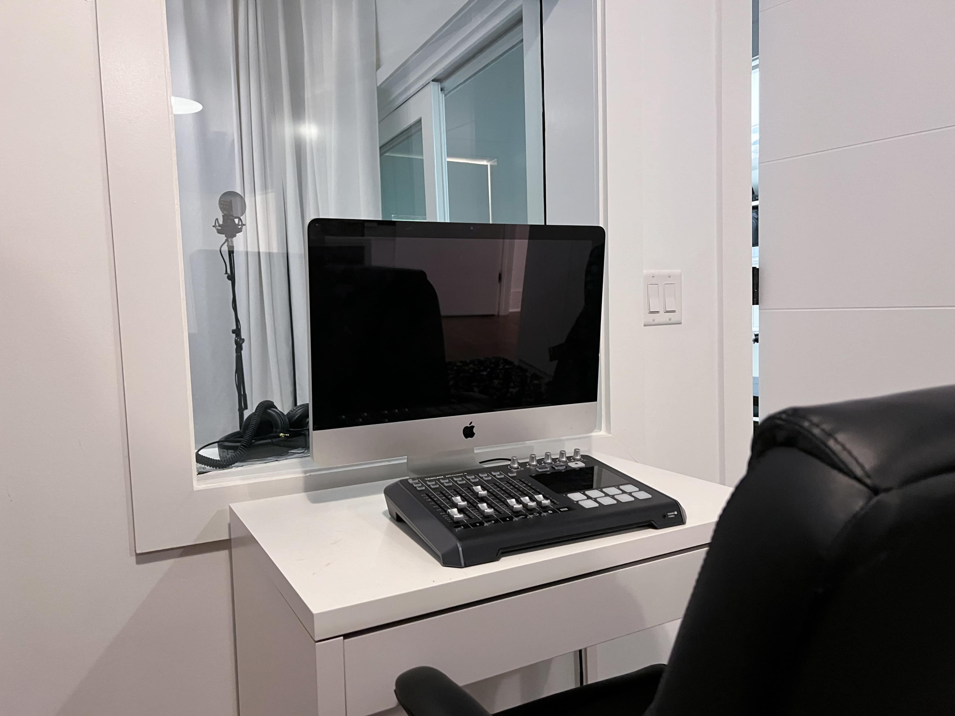A desk setup features an iMac computer and a mixer, with a chair positioned nearby in a well-lit room.