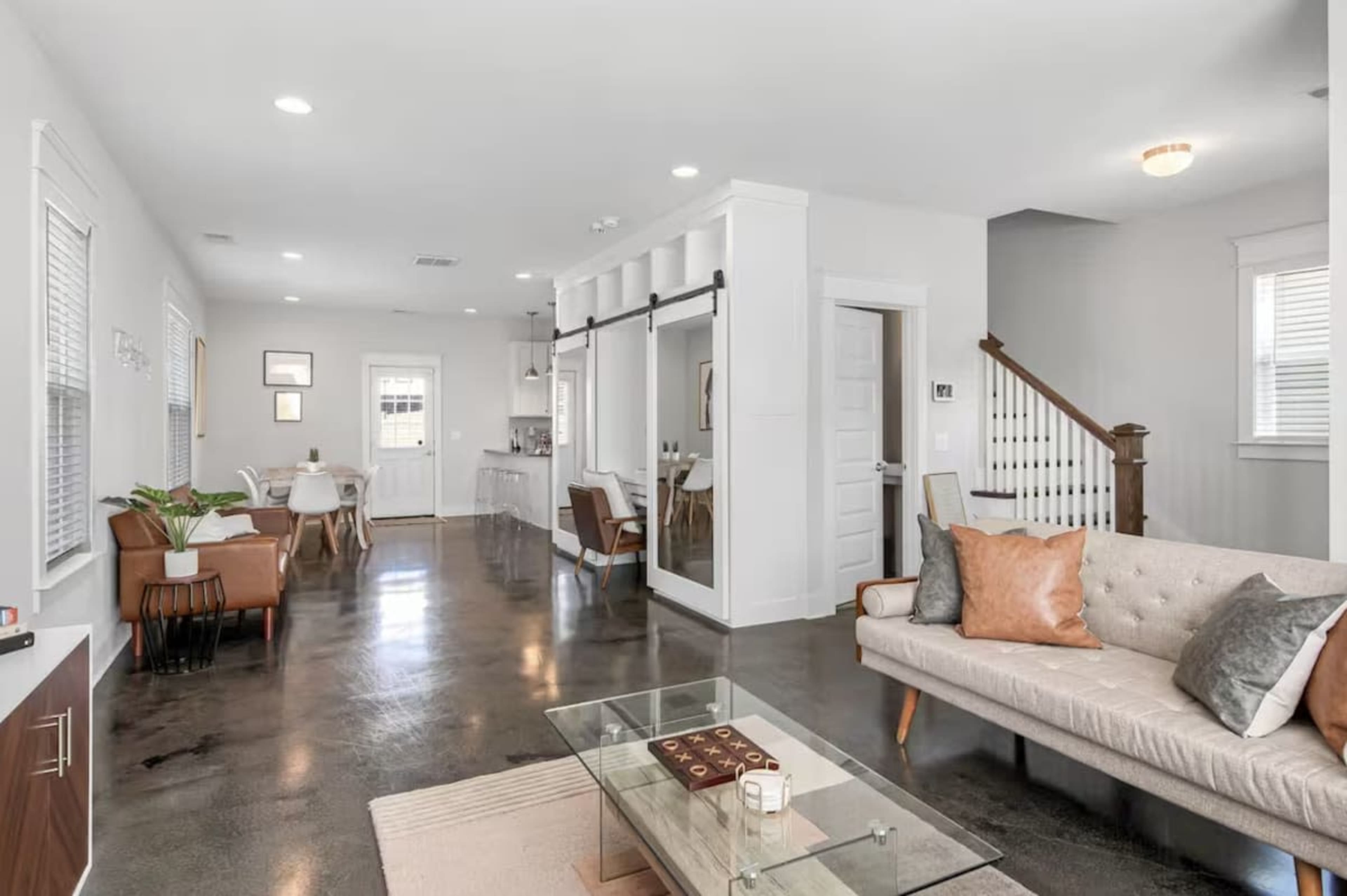 The image shows a modern, open-concept living space featuring a light-colored sofa, a glass coffee table, and a dining area with wooden chairs.